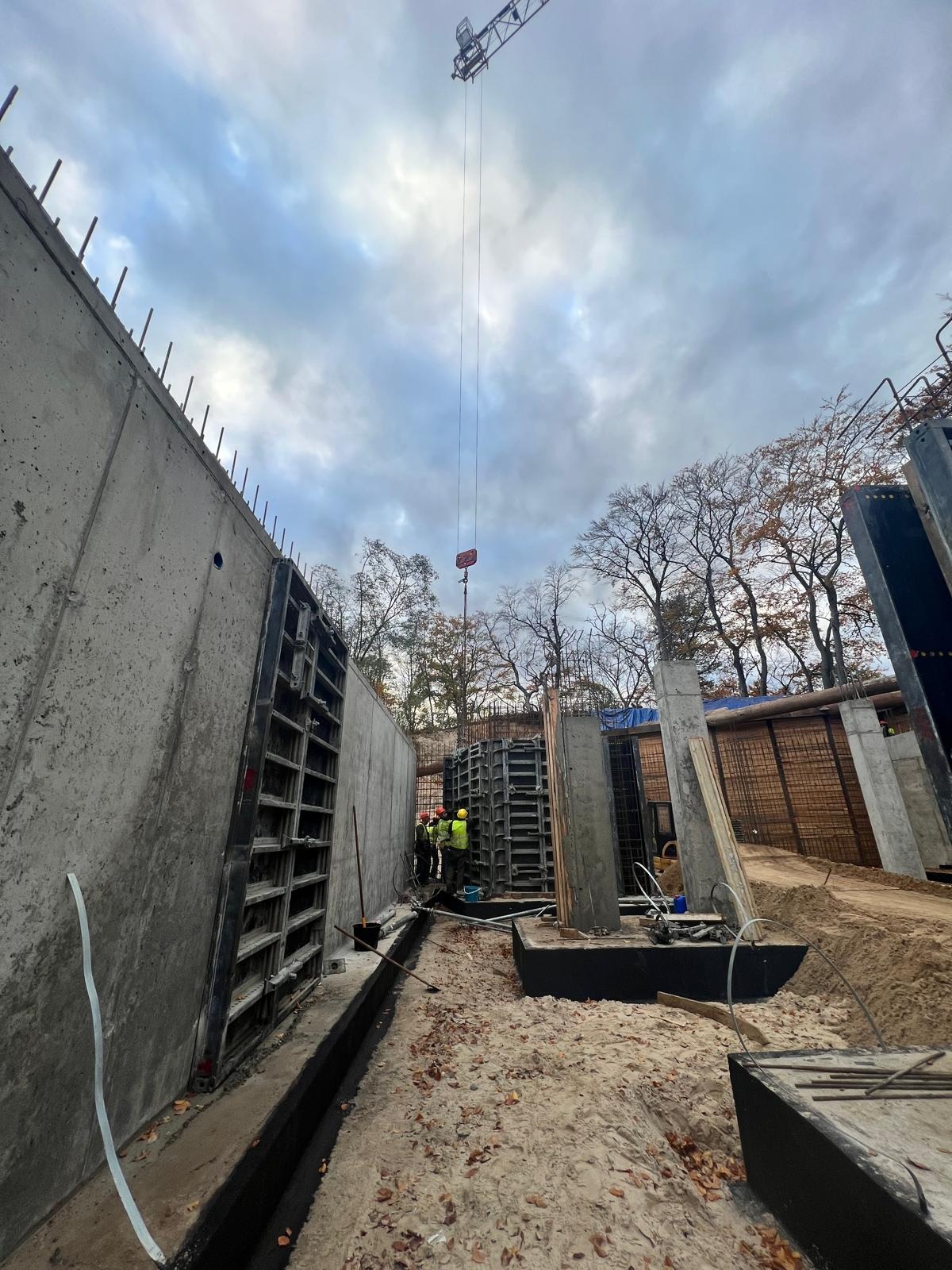 Budowa żelbetowej konstrukcji: widok na szalunki, częściowo wylany beton, zbrojenie i żuraw w tle, jesienne liście na piasku.