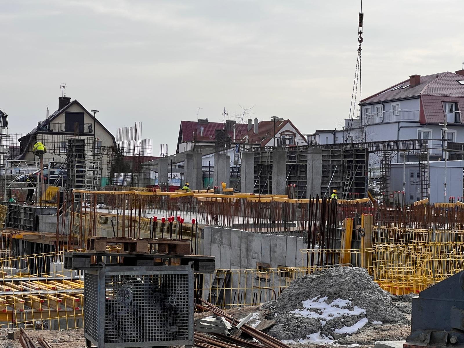 Wznoszenie konstrukcji żelbetowej budynku wielorodzinnego, widoczne szalunki, zbrojenia, pręty, betonowe słupy, pracownicy w kaskach i kamizelkach odblaskowych, dźwig budowlany, w tle zabudowa...