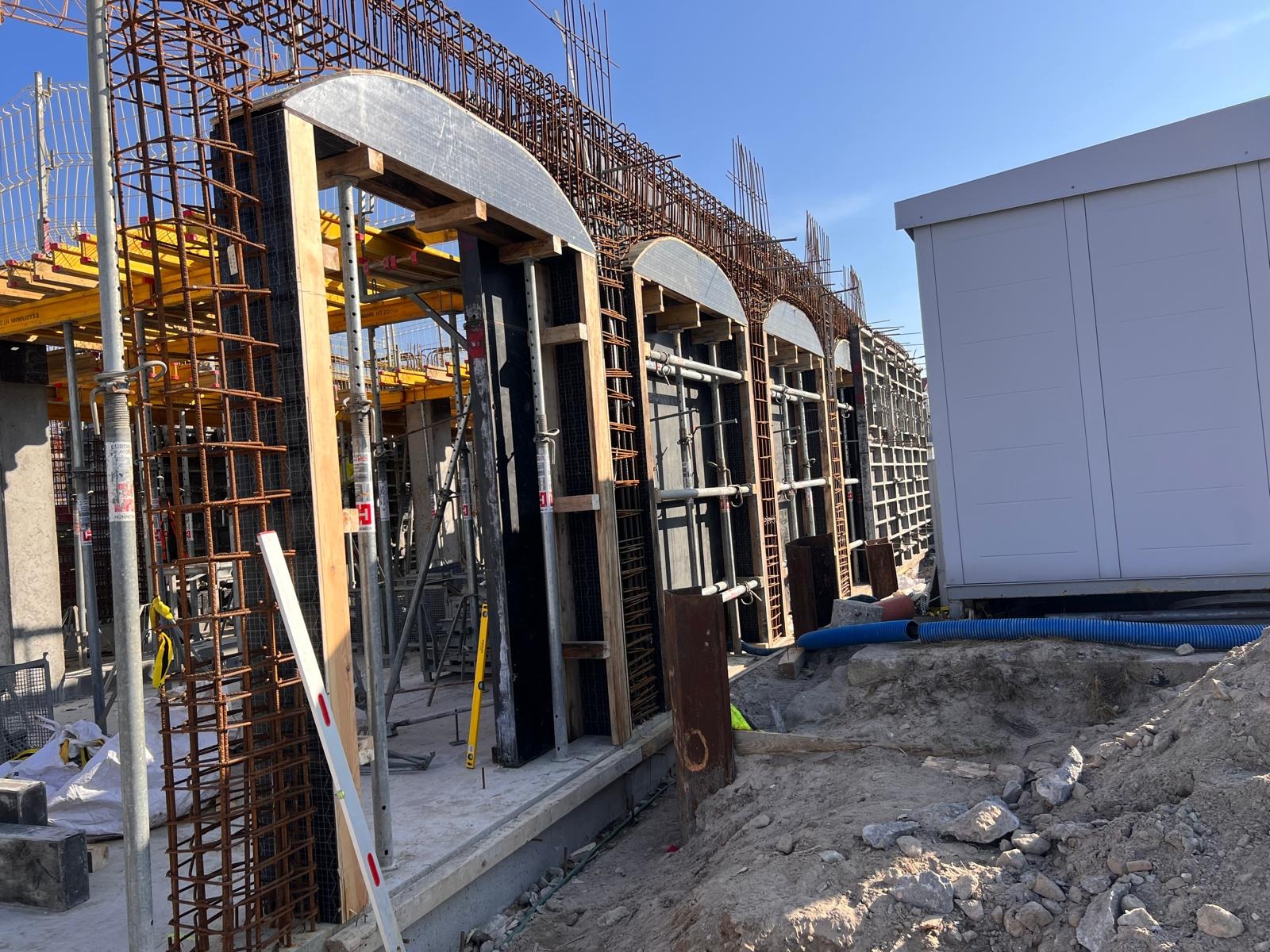 Konstrukcja żelbetowa z szalunkami łukowymi i zbrojeniem stalowym, widoczne podpory i poziomice, obok kontener budowlany, błękitne niebo w tle.