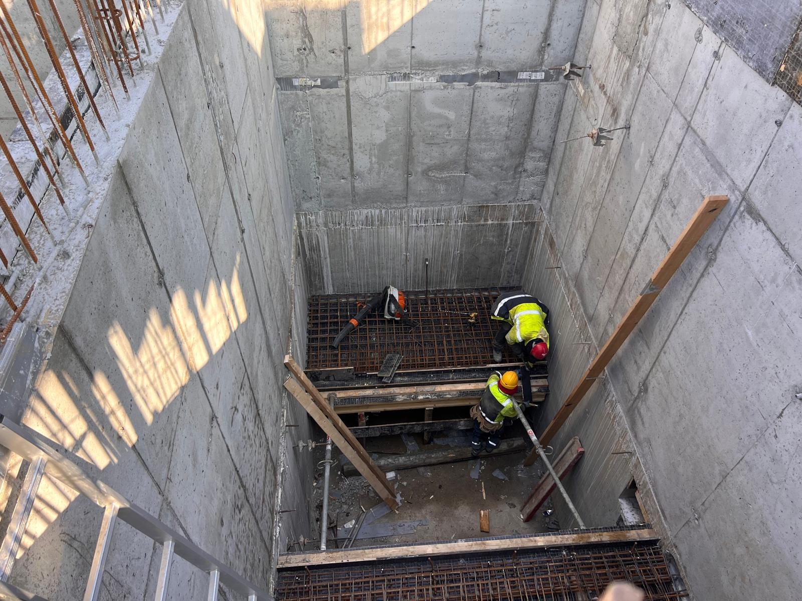 Dwóch pracowników w odblaskowych kamizelkach i kaskach układa zbrojenie wewnątrz betonowego wykopu fundamentowego, widoczna drabina i cień na ścianie.