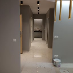 Олександр Даховський - Szary korytarz po malowaniu z marmurową podłogą i złotymi lampami. Widoczne puszki farby i czarne reflektory sufitowe.