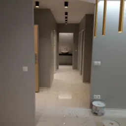 Szary korytarz po malowaniu z marmurową podłogą i złotymi lampami. Widoczne puszki farby i czarne reflektory sufitowe.