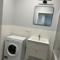 Олександр Даховський - Nowoczesna łazienka w szarościach: pralka, umywalka z szafką w geometryczny wzór, lustro z oświetleniem. Widoczne płytki i armatura. Minimalistyczny design.