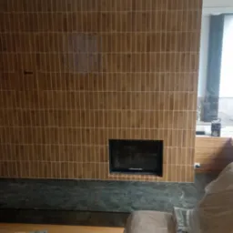 Wykończony kominek z brązową, pionowo ułożoną, wąską glazurą i ciemnozielonym marmurem. Widoczne duże okno i elementy wyposażenia wnętrza w trakcie prac.