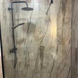 Kabina prysznicowa z deszczownicą i szklaną ścianką, wykończona płytkami imitującymi marmur w odcieniach szarości i beżu. Nowoczesny design, minimalistyczne wykończenie.