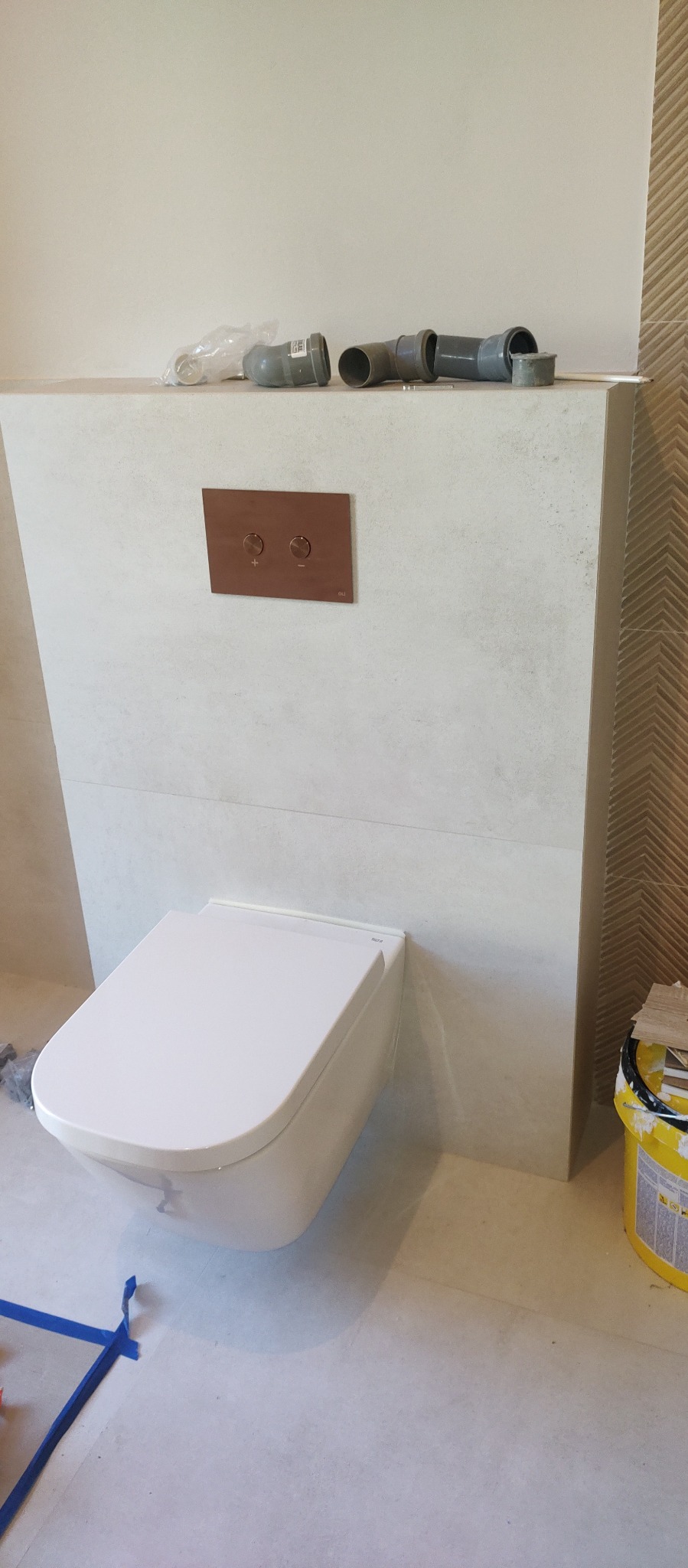 Zamontowana, podwieszana toaleta z przyciskiem w kolorze miedzi na tle jasnych płytek. Częściowo widoczne rury i narzędzia. W tle dekoracyjna ściana.