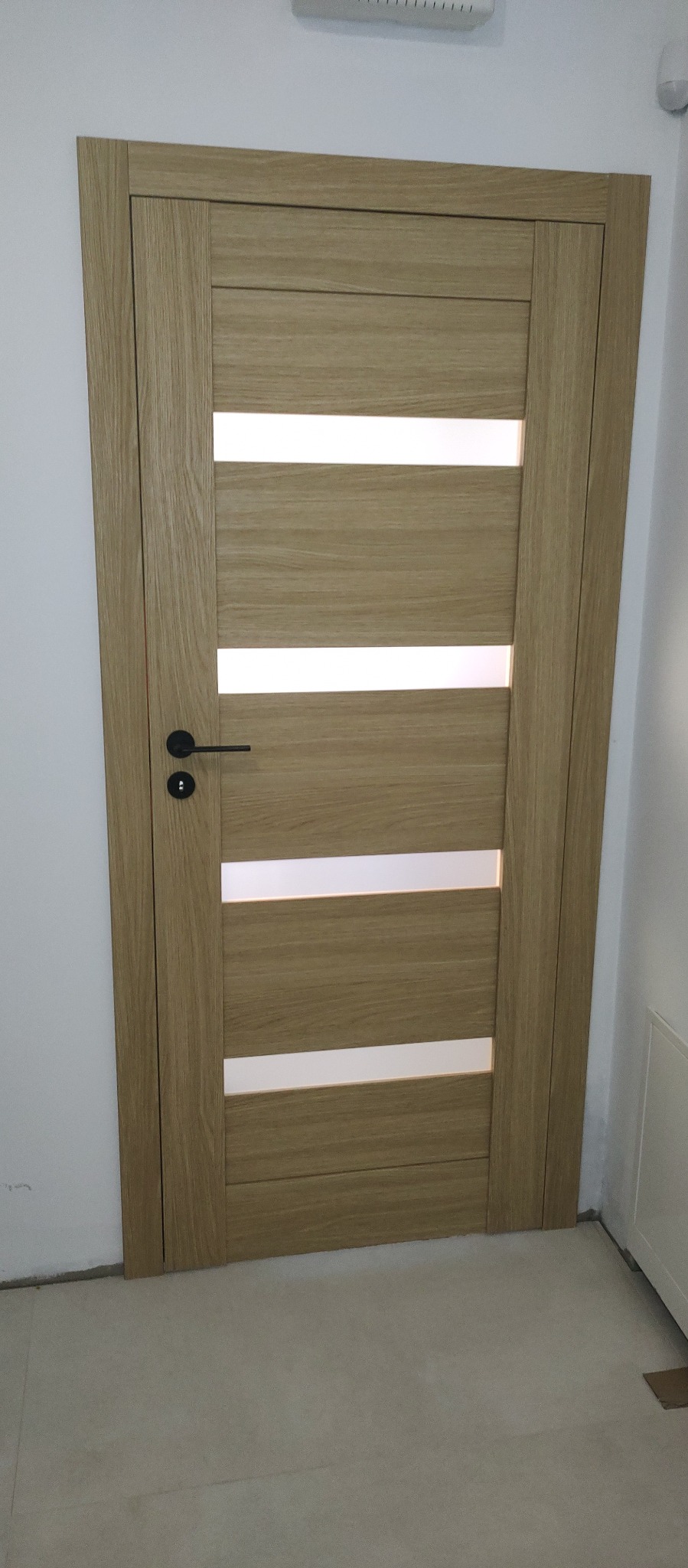 Jasne, drewniane drzwi wewnętrzne z poziomymi, matowymi przeszkleniami i czarną klamką, zamontowane w białej ścianie. Widoczny fragment podłogi.