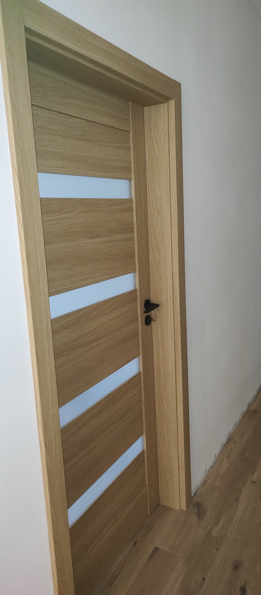 Jasne, drewniane drzwi wewnętrzne z czterema poziomymi, mlecznymi przeszkleniami i czarną klamką, zamontowane w białej ścianie na tle drewnianej podłogi.