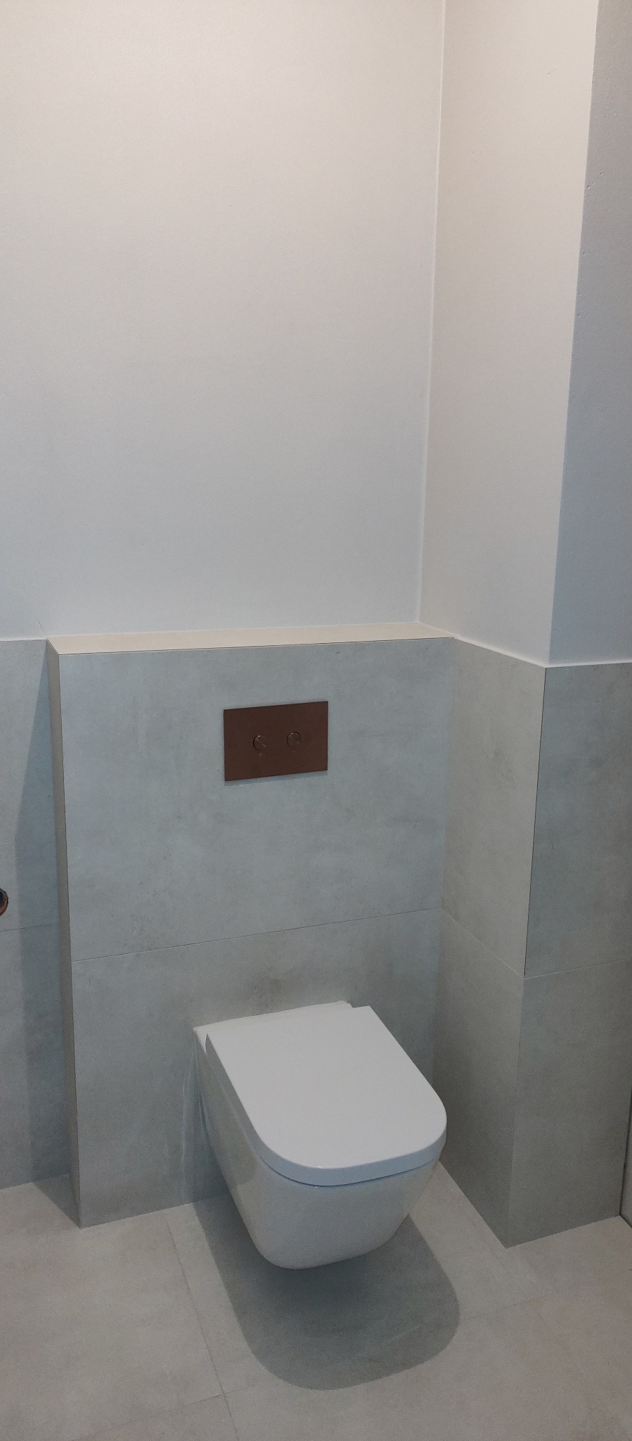 Nowoczesna, podwieszana toaleta z brązowym panelem spłukującym na tle szarych, matowych płytek w wykończonej łazience. Minimalistyczny design.