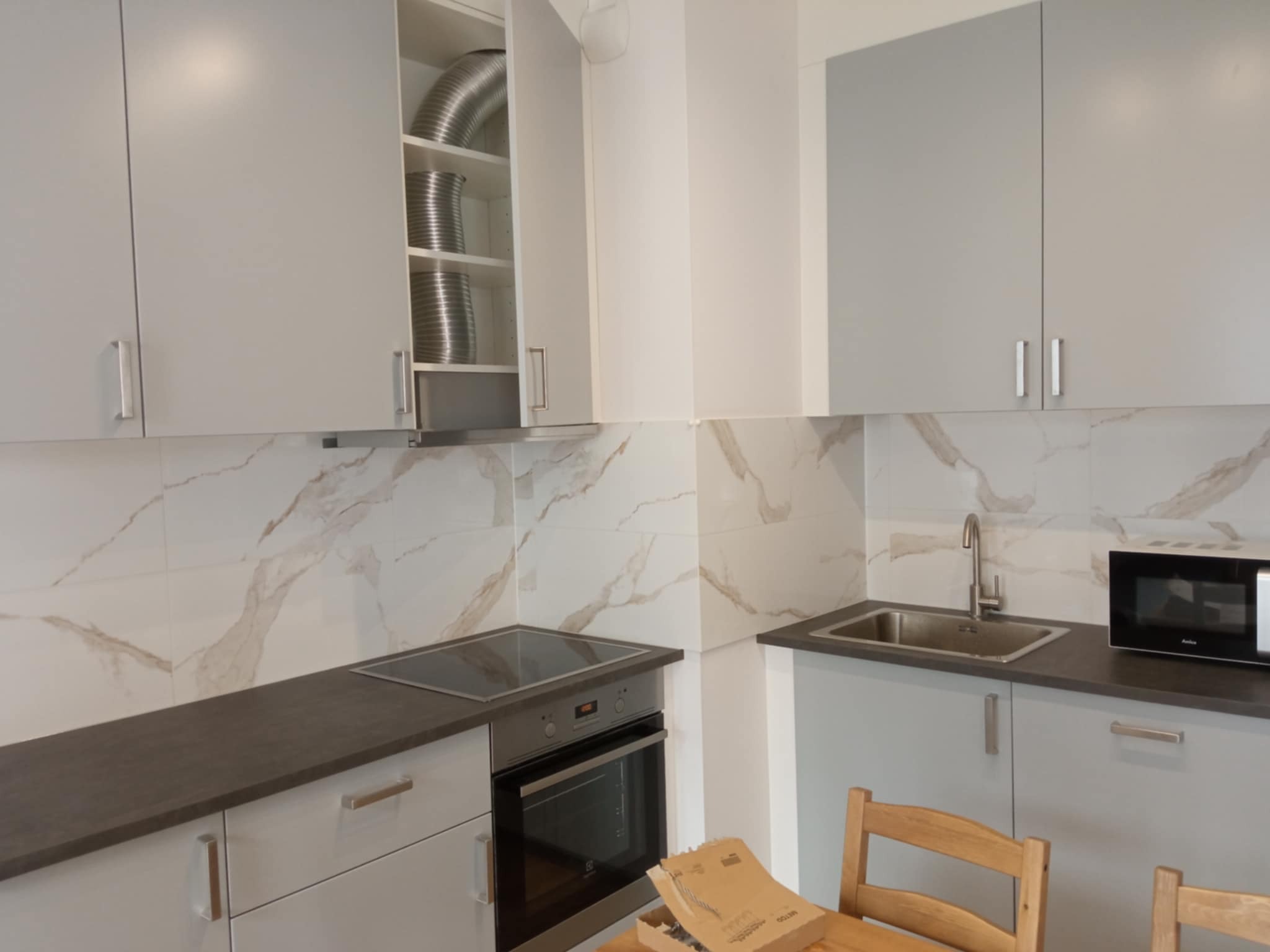 Jasna kuchnia po remoncie: białe szafki, marmurowe płytki na ścianie, szary blat. W otwartej szafce widoczna metalowa rura wentylacyjna.