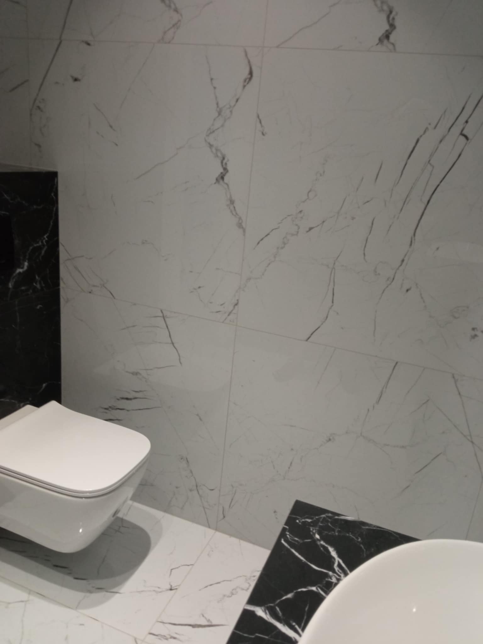 Wykończona łazienka z białymi płytkami imitującymi marmur, kontrastującymi z czarnymi elementami. Uwagę przykuwa podwieszana toaleta i fragment umywalki.