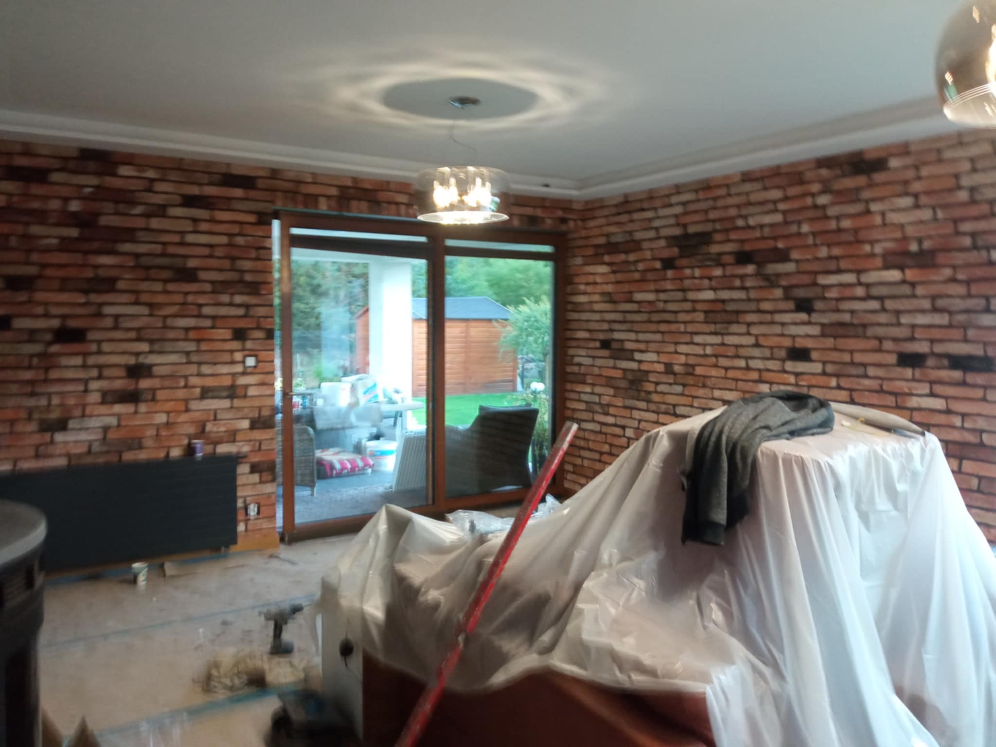 Ceglana ściana w pokoju z dużym oknem, widoczne zabezpieczone folią meble i narzędzia. Wnętrze w trakcie prac wykończeniowych, lampa sufitowa.