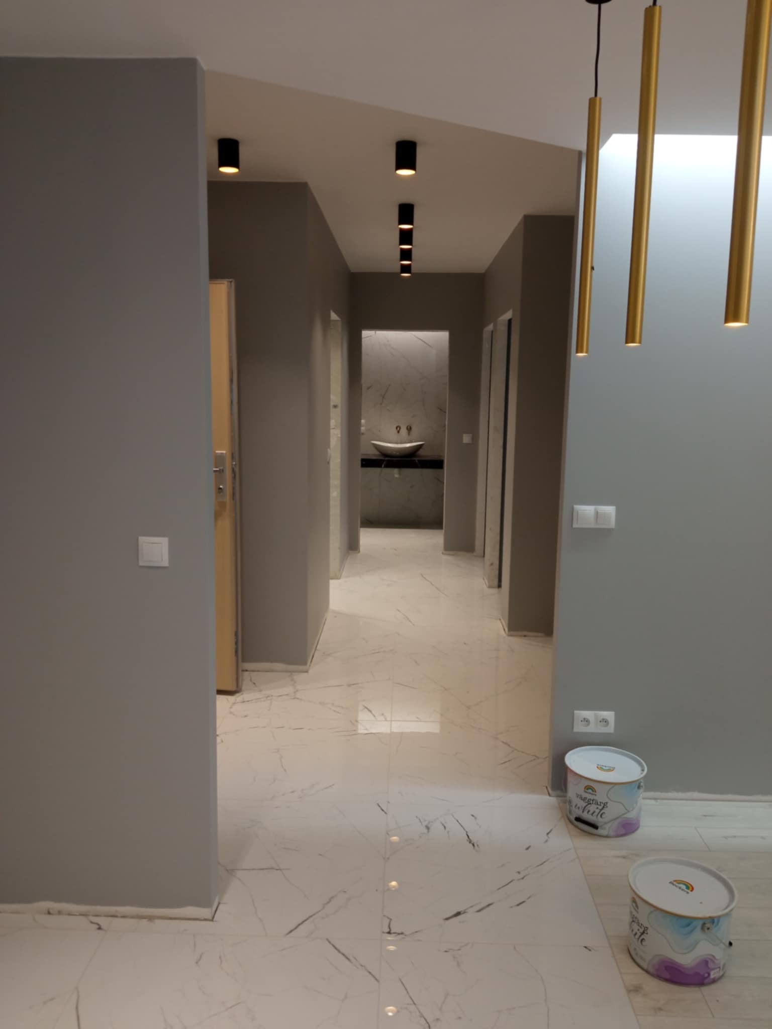 Szary korytarz po malowaniu z marmurową podłogą i złotymi lampami. Widoczne puszki farby i czarne reflektory sufitowe.