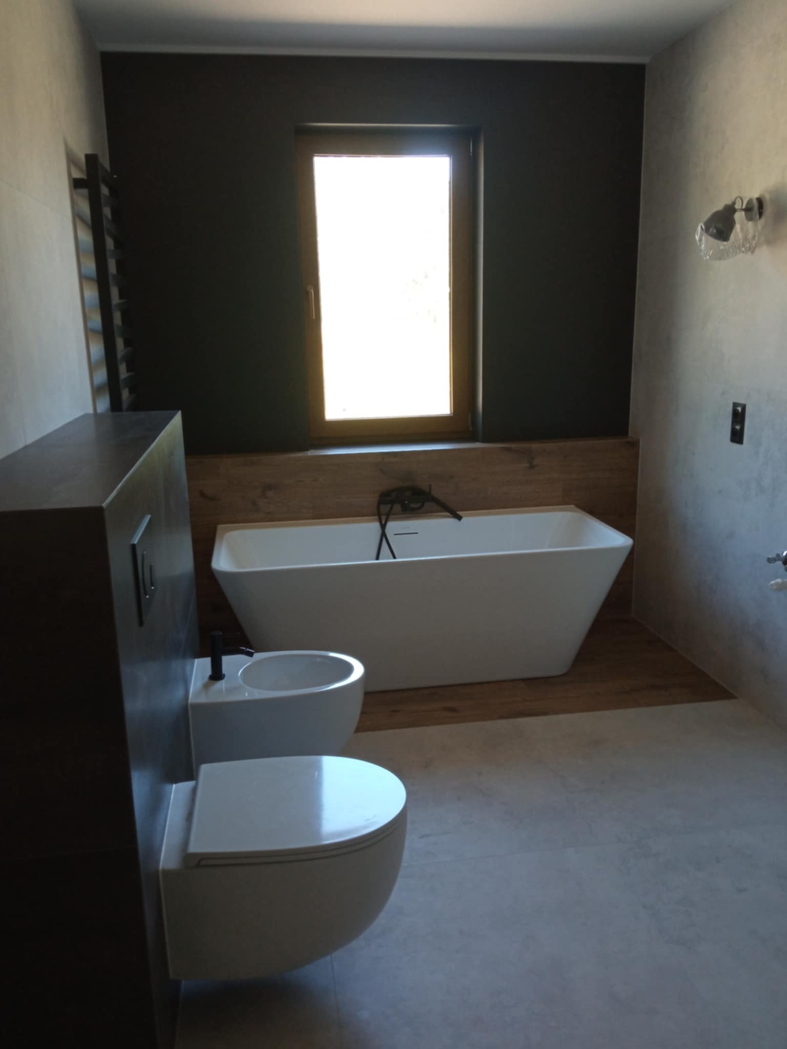 Nowoczesna łazienka z wolnostojącą wanną, bidetem i podwieszaną toaletą. Ciemna ściana kontrastuje z drewnianym wykończeniem wnęki wanny i jasnymi płytkami.