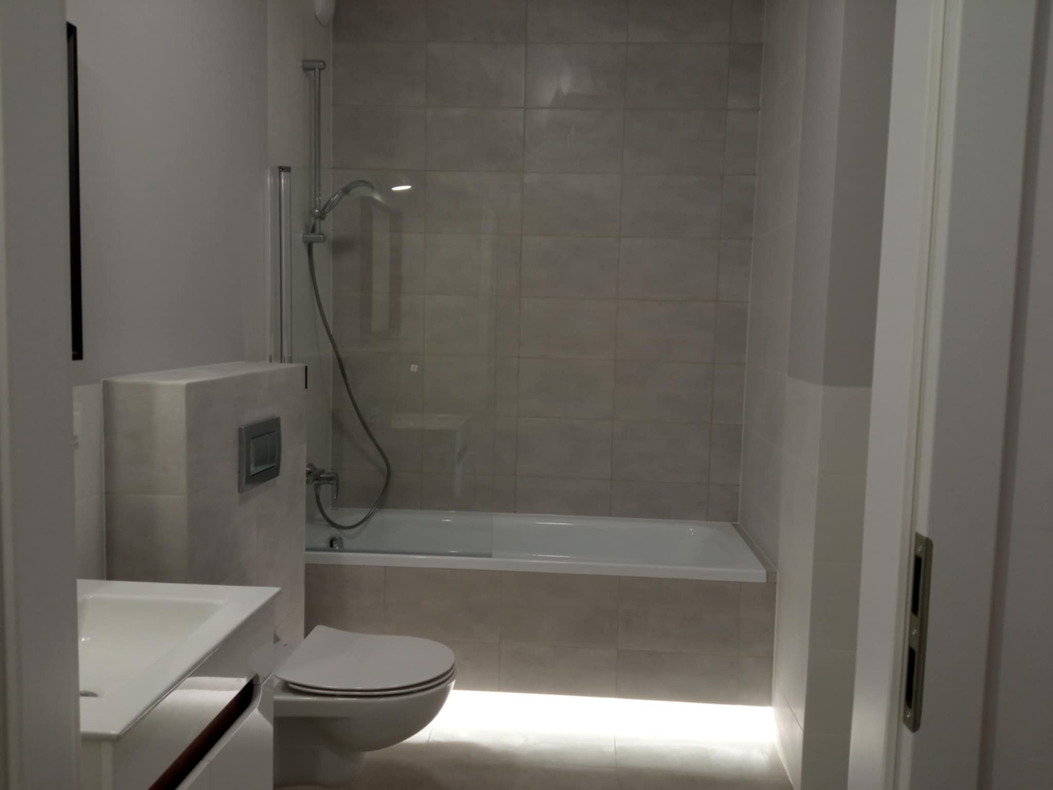 Jasna łazienka z wanną za szklaną ścianką, umywalką z szafką i podświetlaną podłogą. Minimalistyczny design, neutralne kolory, nowoczesne wykończenie.