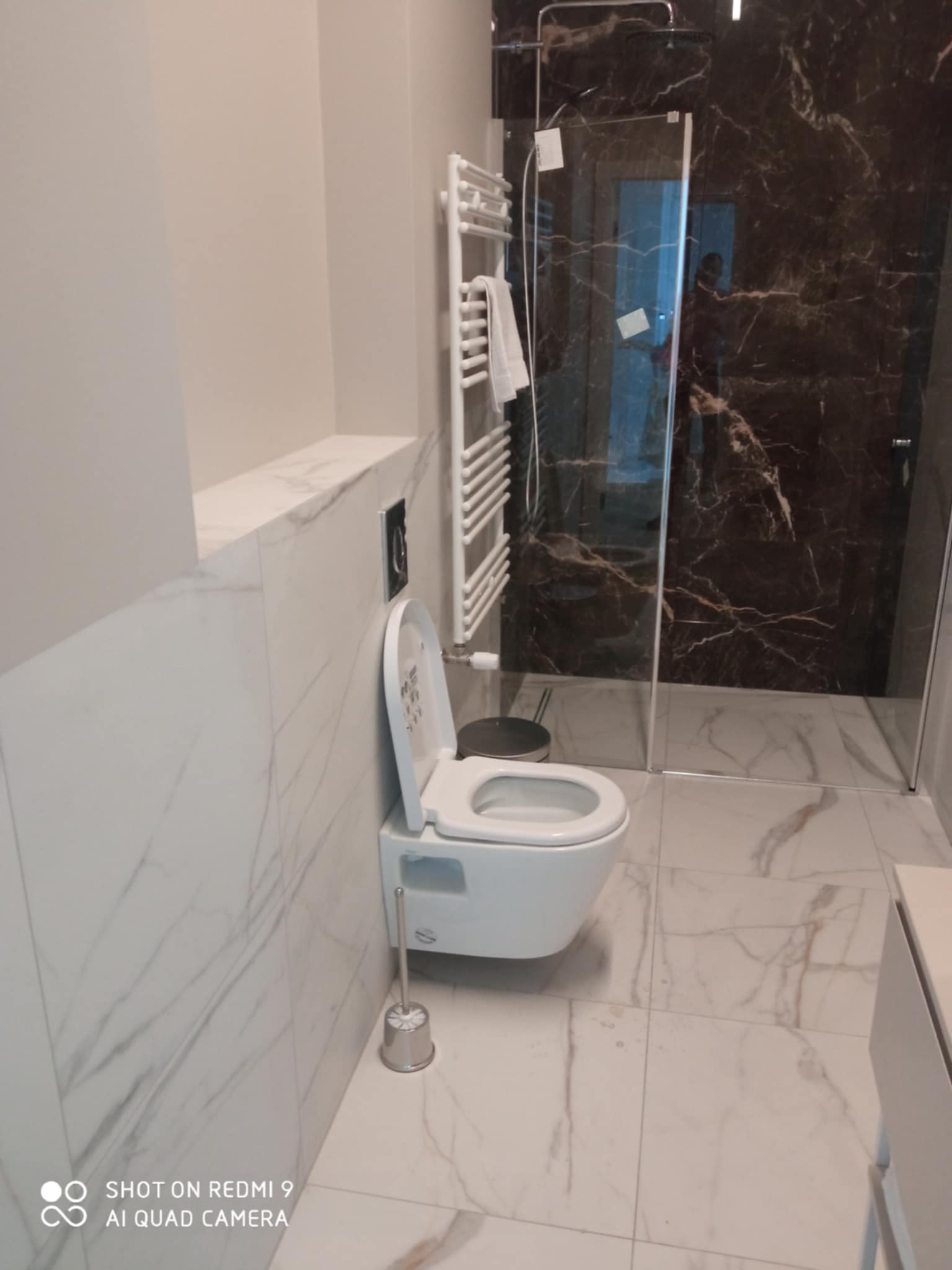 Nowoczesna łazienka z marmurowymi płytkami, podwieszaną toaletą i kabiną prysznicową z ciemnego marmuru. Biały grzejnik drabinkowy z ręcznikiem.