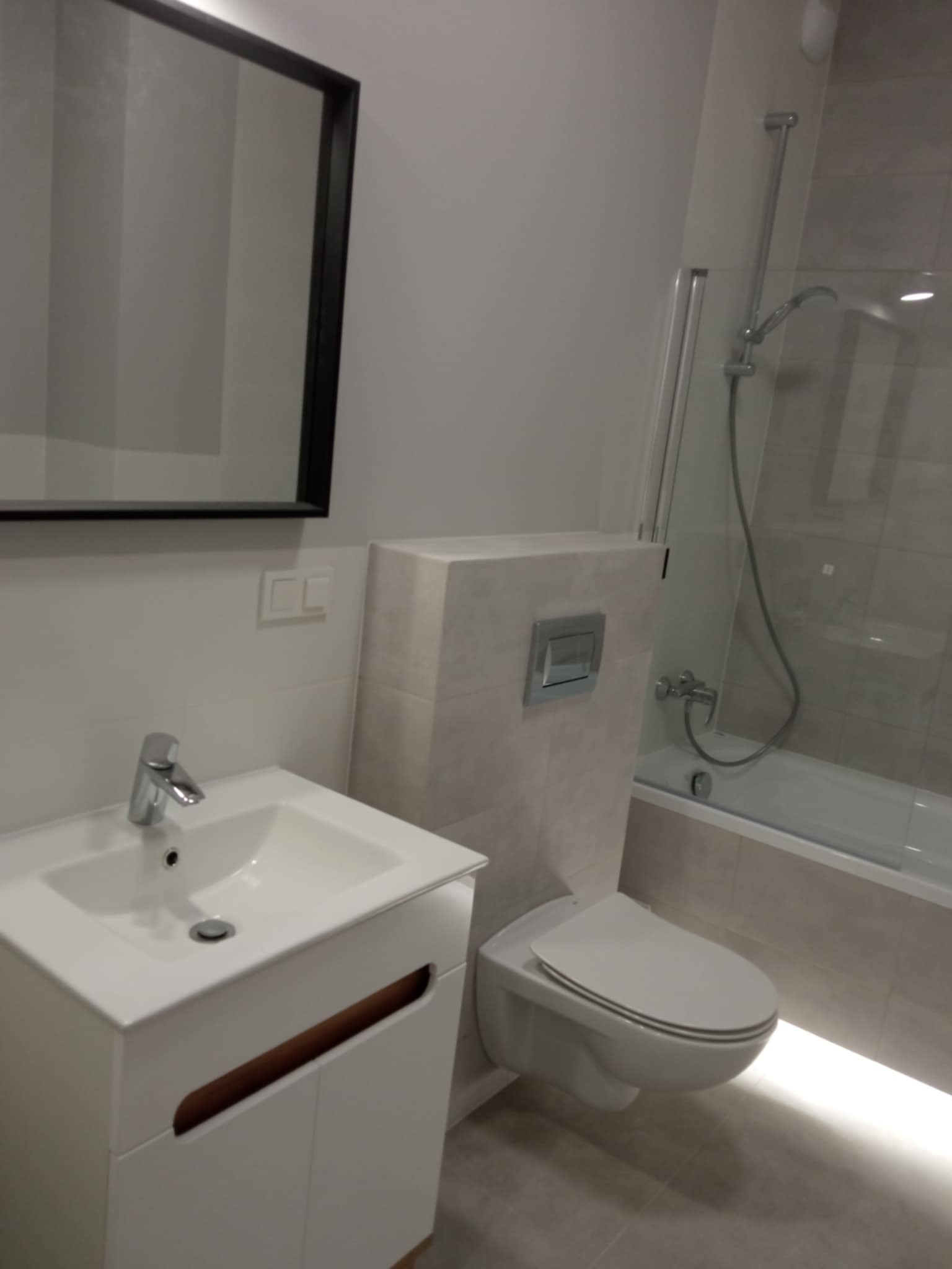 Nowoczesna łazienka po remoncie: biała umywalka z szafką, podwieszana toaleta, lustro w czarnej ramie, prysznic za szklaną ścianką i podświetlenie LED.