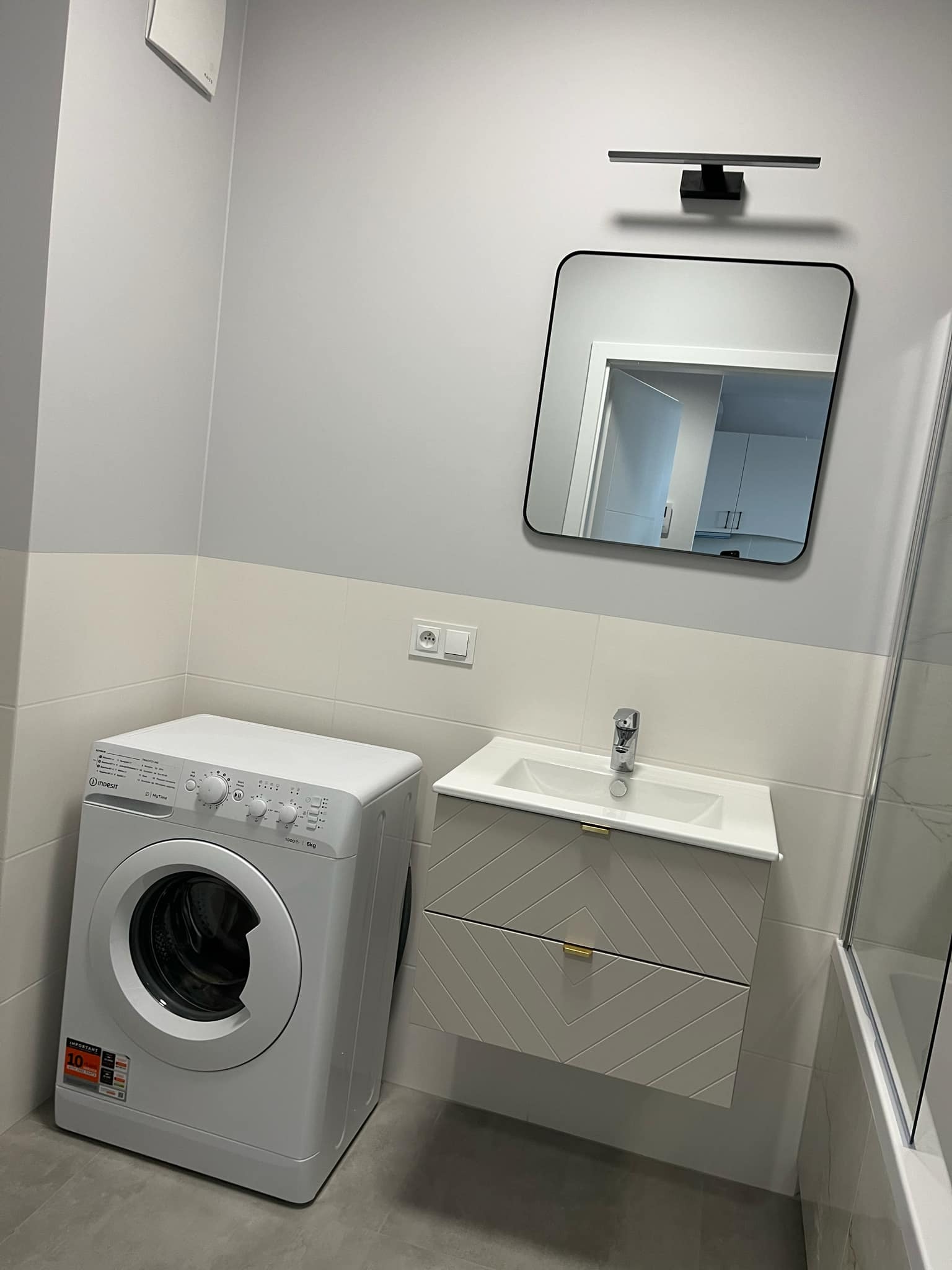 Nowoczesna łazienka w szarościach: pralka, umywalka z szafką w geometryczny wzór, lustro z oświetleniem. Widoczne płytki i armatura. Minimalistyczny design.