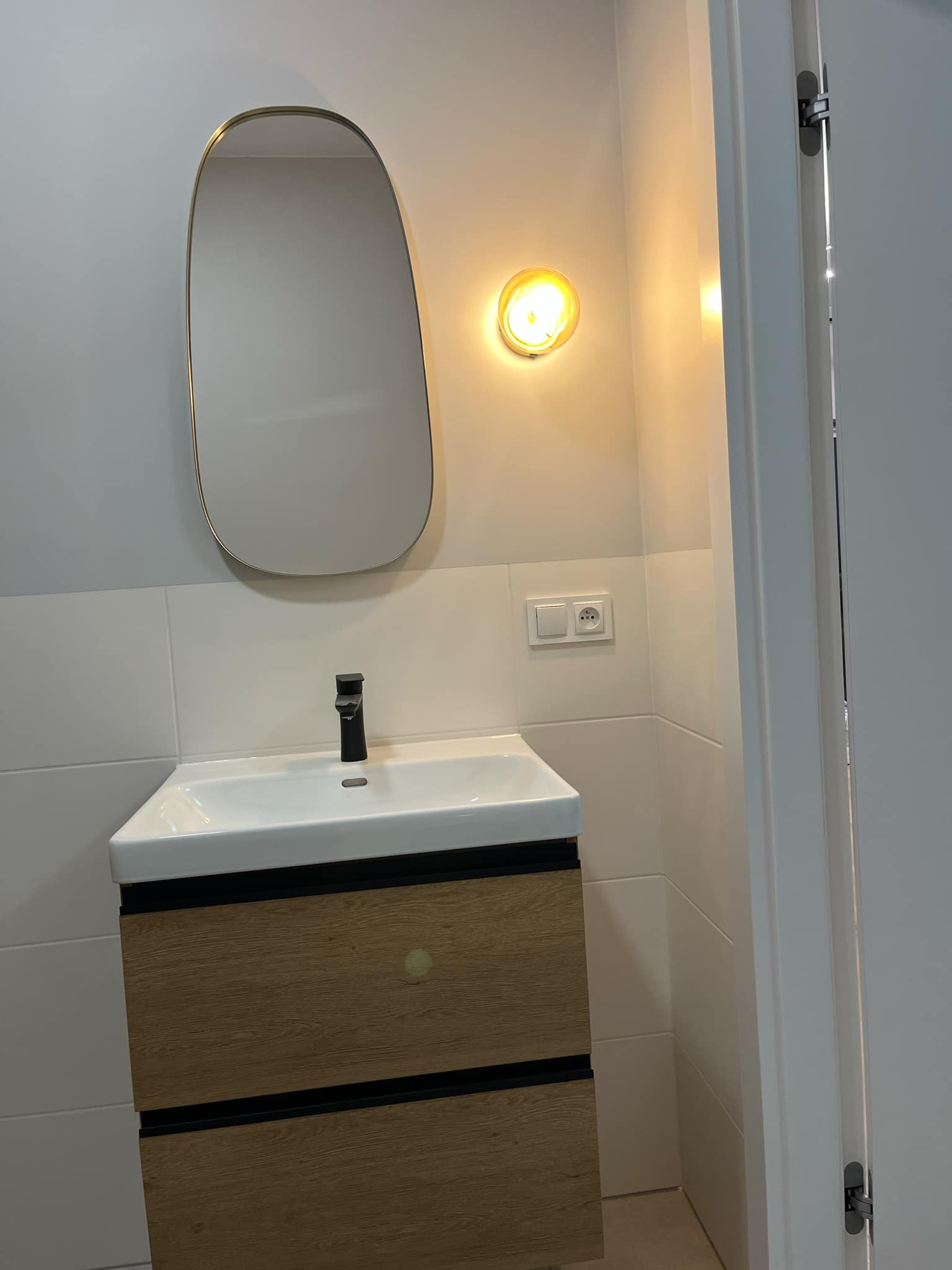 Nowoczesna łazienka w Grójcu: białe płytki, szafka z drewnianym frontem, asymetryczne lustro i okrągła lampa nad umywalką. Minimalistyczny design.