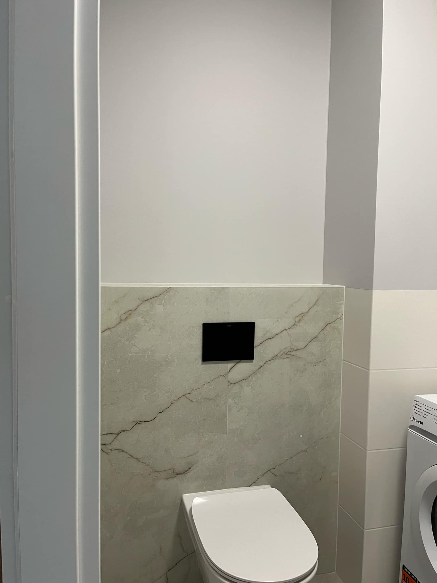 Wnętrze łazienki po remoncie: podwieszana toaleta z marmurową płytką, czarny panel sterujący, pralka Indesit. Estetyczne wykończenie.