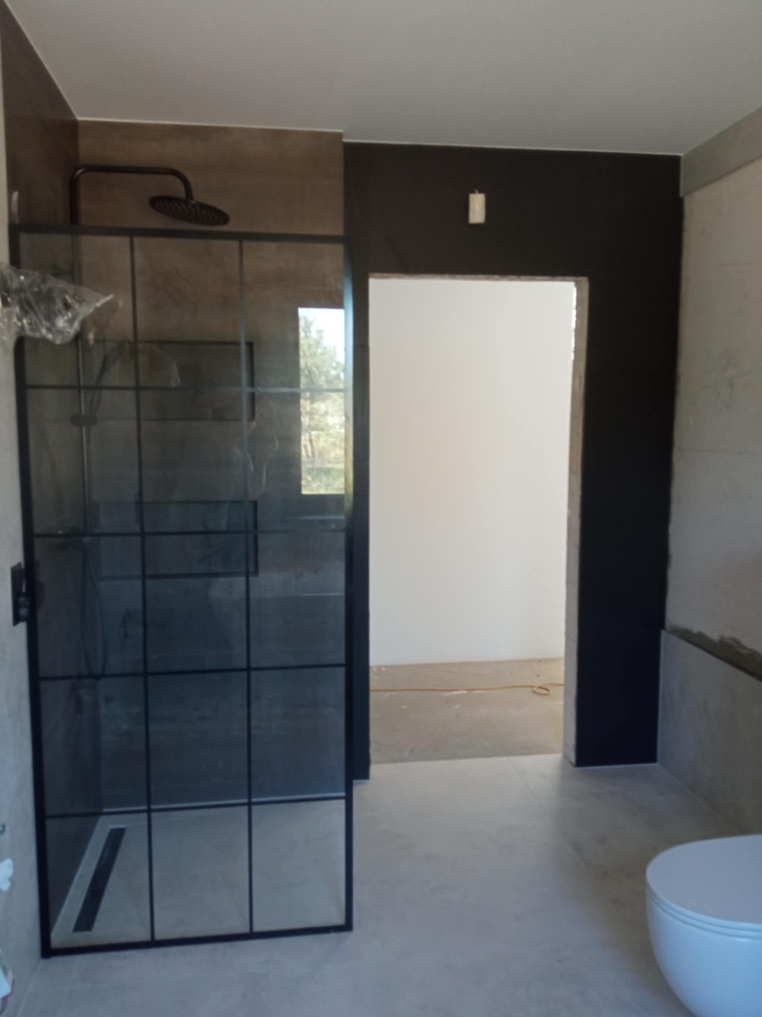 Nowoczesna kabina prysznicowa z czarnymi profilami i deszczownicą w wykończonej łazience z szarą glazurą i czarną ścianą. Widok na sąsiednie pomieszczenie.