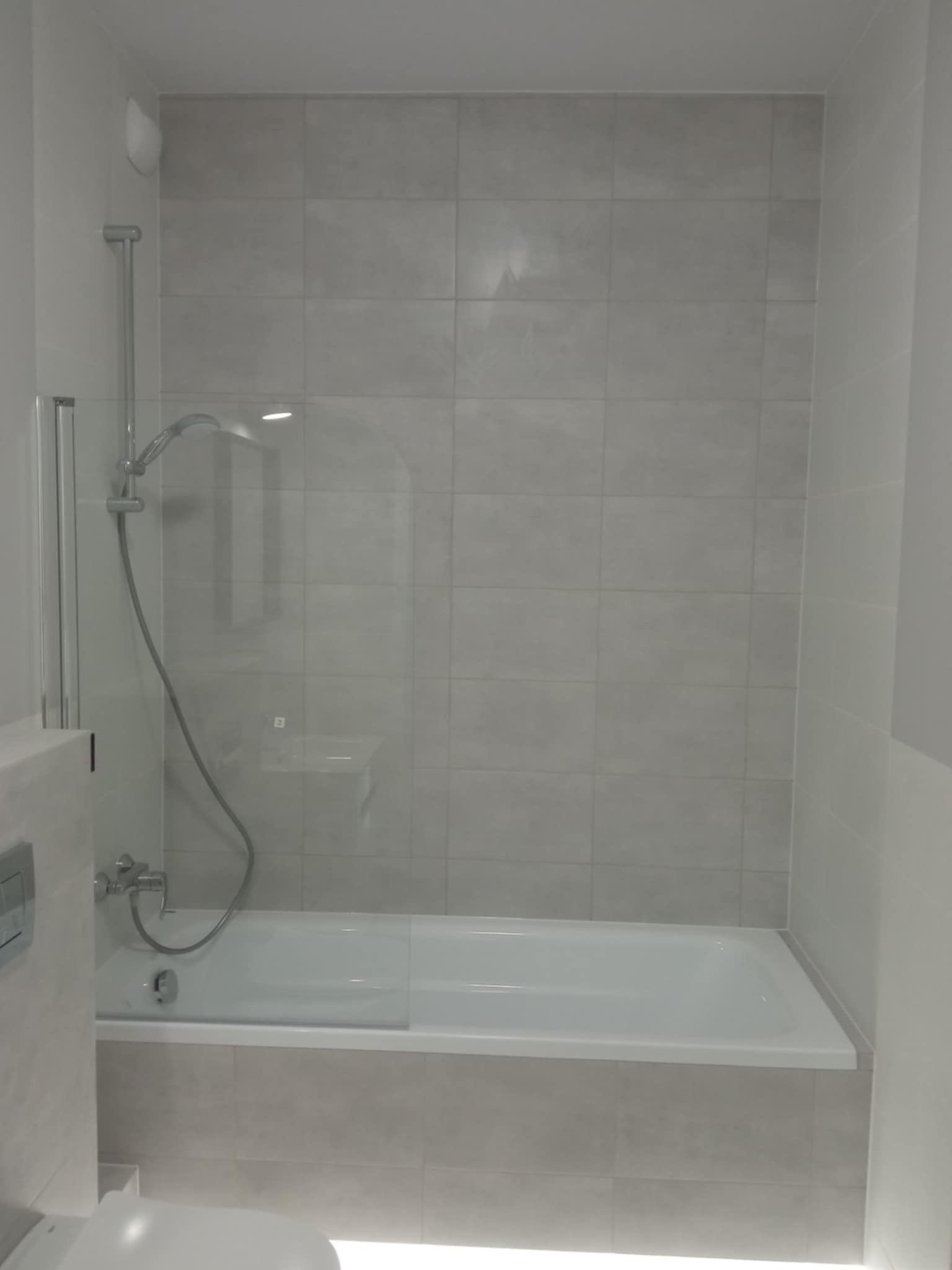 Jasna łazienka z wanną i szklanym parawanem nawannowym, wykończona w szarych płytkach. Nowoczesna armatura i minimalistyczny design.