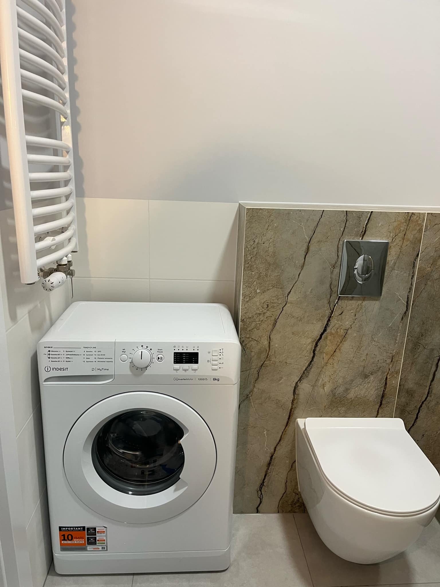 Wykończona łazienka: biała pralka Indesit obok podwieszanej toalety, grzejnik drabinkowy na ścianie z jasnymi płytkami, akcenty marmurowe. Nowoczesny design.