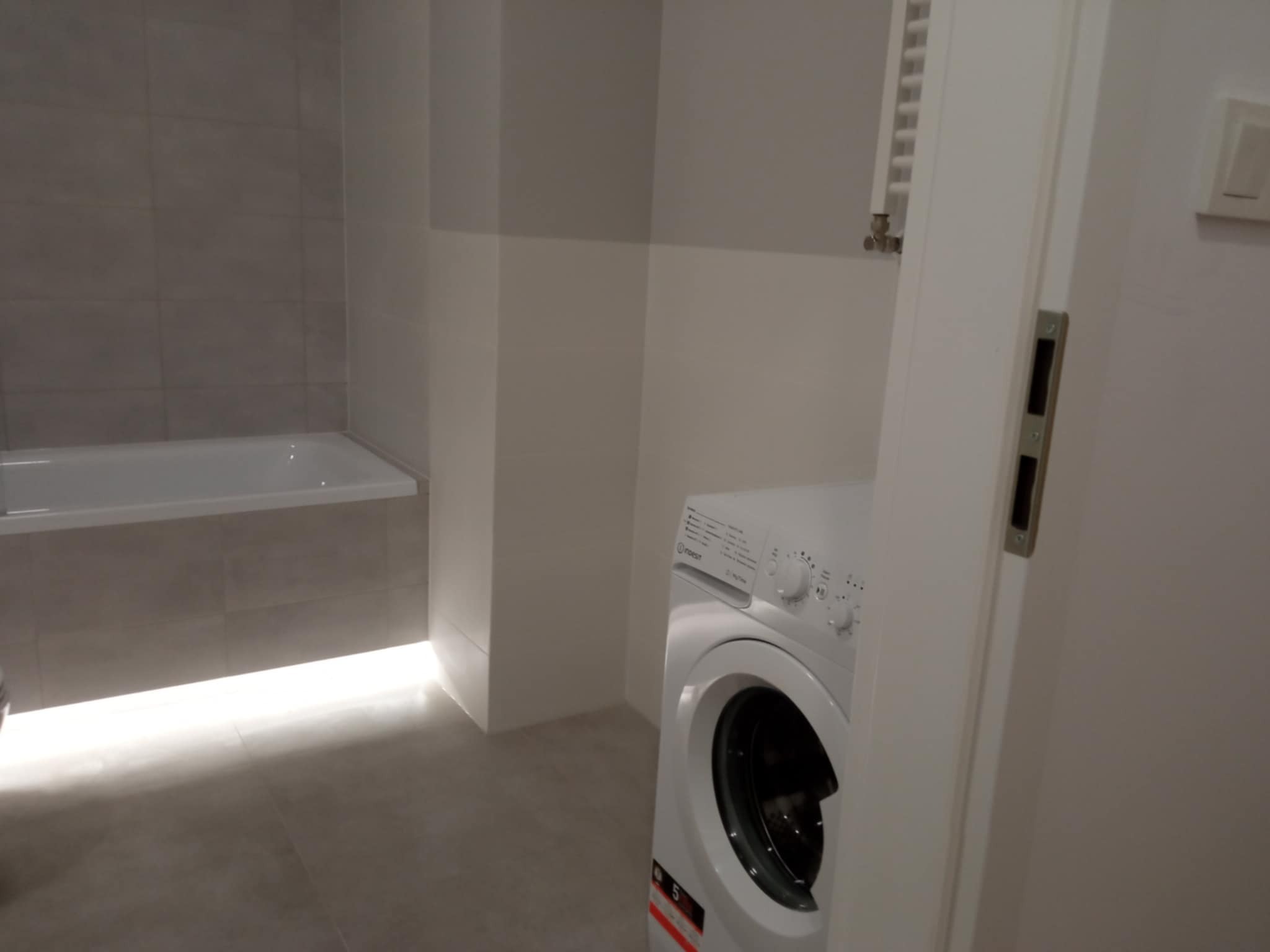 Wykończona łazienka z podświetlaną wanną, szarymi płytkami i białą pralką Inodesit, widoczna zza uchylonych drzwi. Nowoczesny design, neutralna kolorystyka.