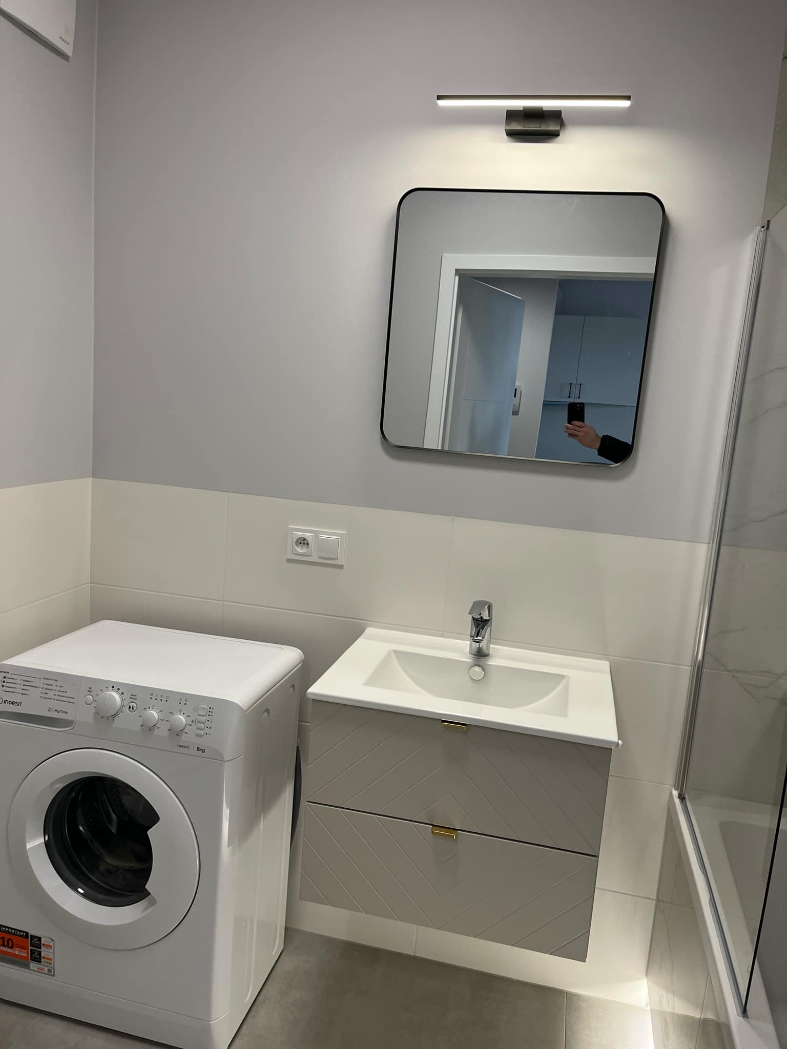 Nowoczesna łazienka z pralką, umywalką z szafką w odcieniach szarości i lustrem. Minimalistyczne oświetlenie i płytki w dwóch odcieniach szarości.
