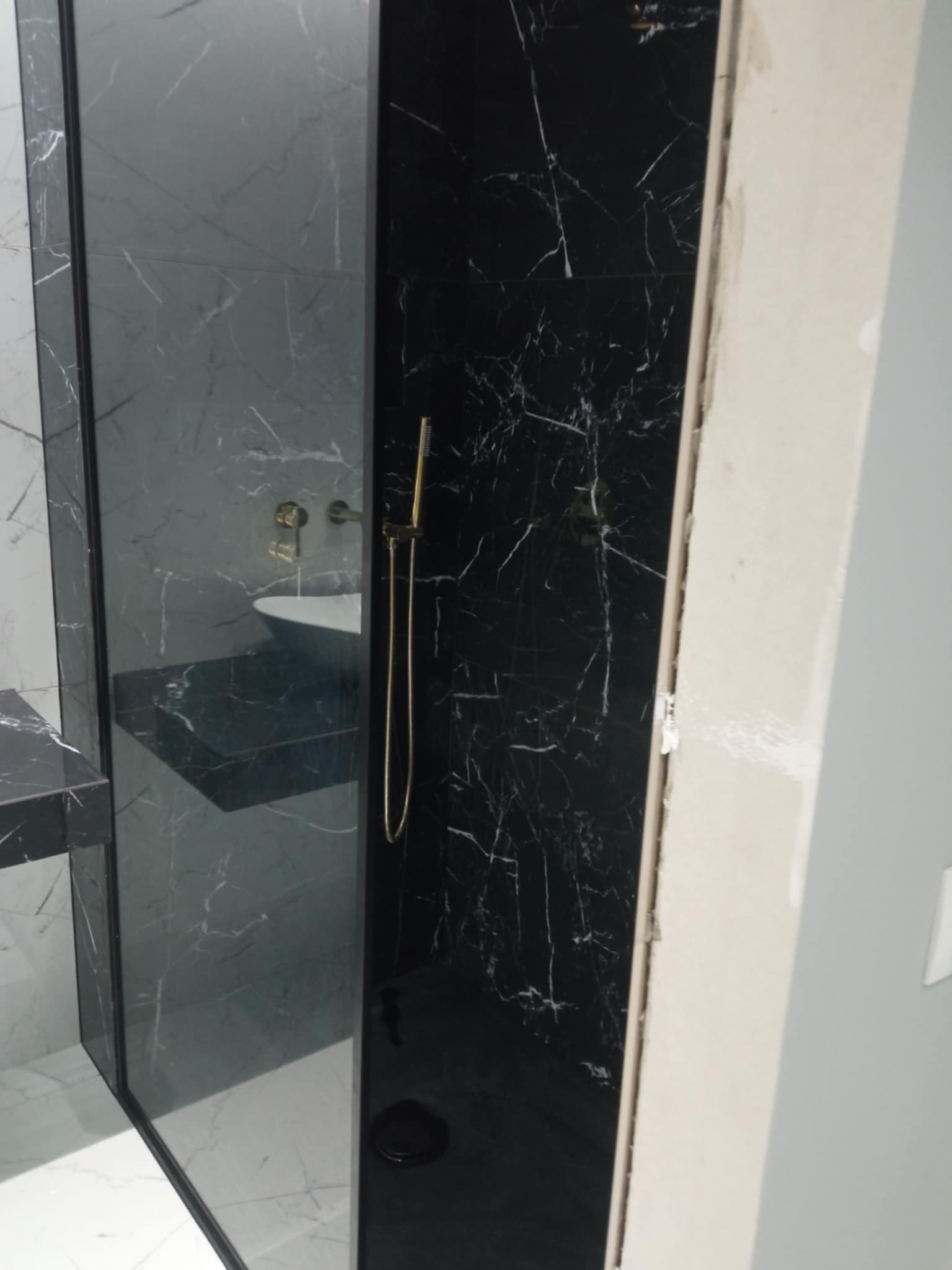 Wykończona kabina prysznicowa z czarnymi i szarymi marmurowymi płytkami, złotą armaturą. Widoczny fragment umywalki i niedokończone obramowanie drzwi.