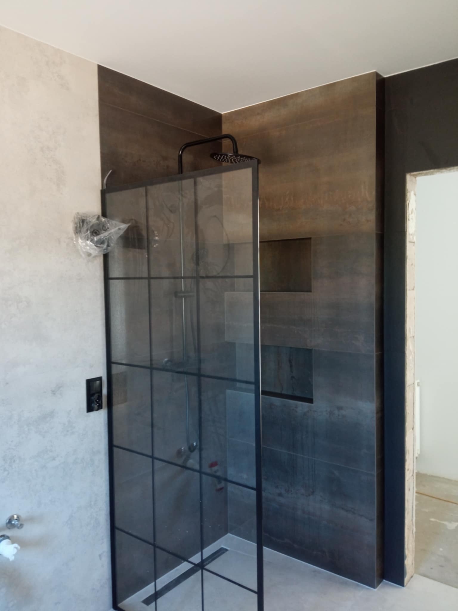 Nowoczesna kabina prysznicowa z czarną ramą i deszczownicą, wykończona płytkami w odcieniach szarości i brązu. Widoczne elementy instalacji w trakcie montażu.