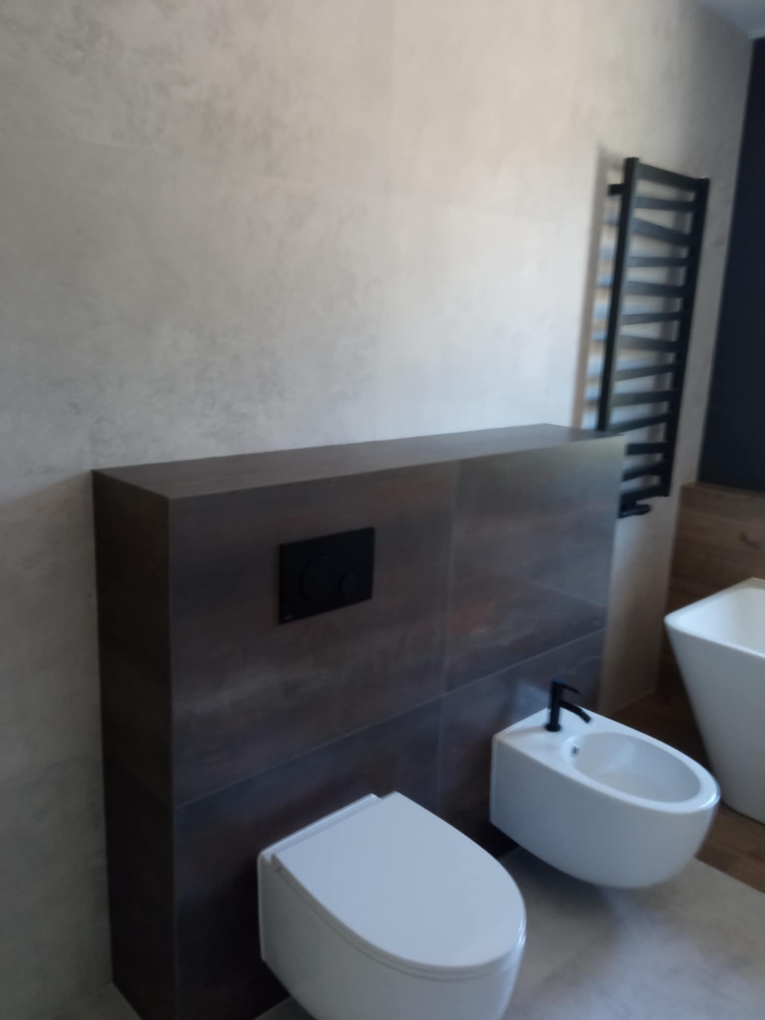 Nowoczesna łazienka: podwieszana toaleta i bidet na tle ściany z płytek w odcieniach brązu i beżu, z grzejnikiem drabinkowym w tle. Minimalistyczny design.