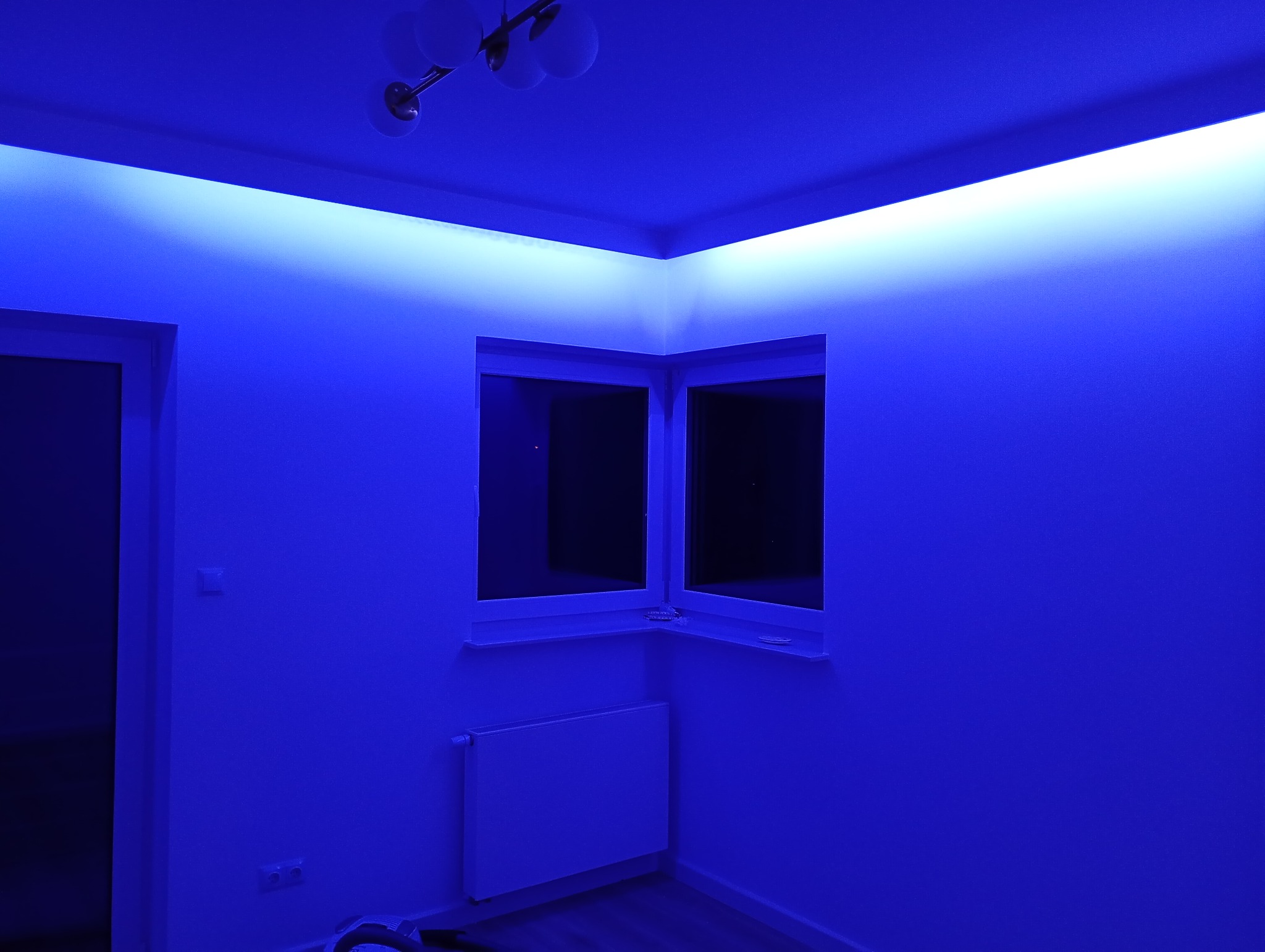 Pokój z oświetleniem LED w kolorze niebieskim, widoczne okno narożne, grzejnik i lampa sufitowa. Nowoczesne wykończenie wnętrza.