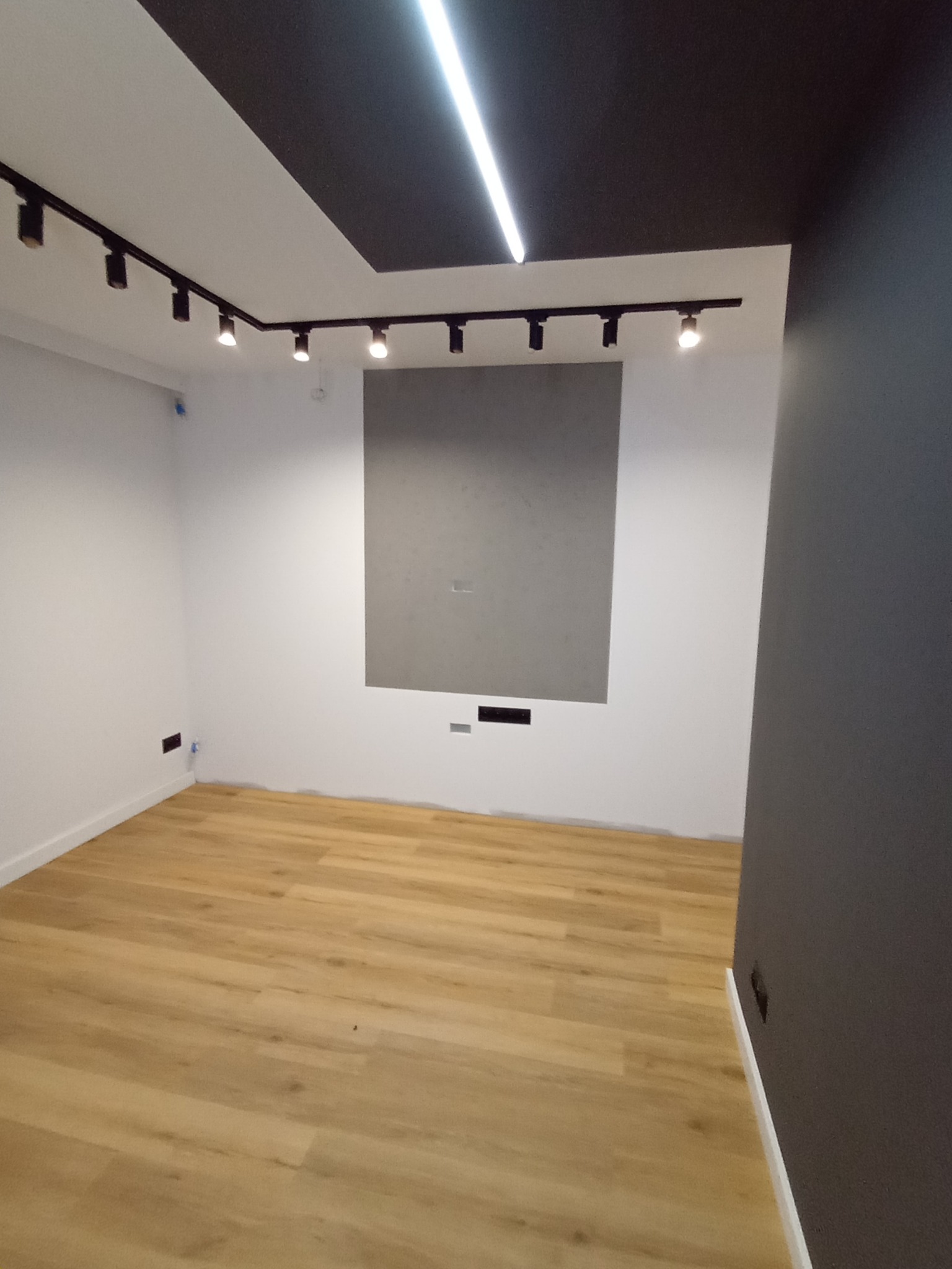 Jasne panele podłogowe w pokoju z białymi ścianami i szarą wstawką. Na suficie czarne szyny z reflektorami i biała lampa liniowa. Ściana po prawej pomalowana na szaro.