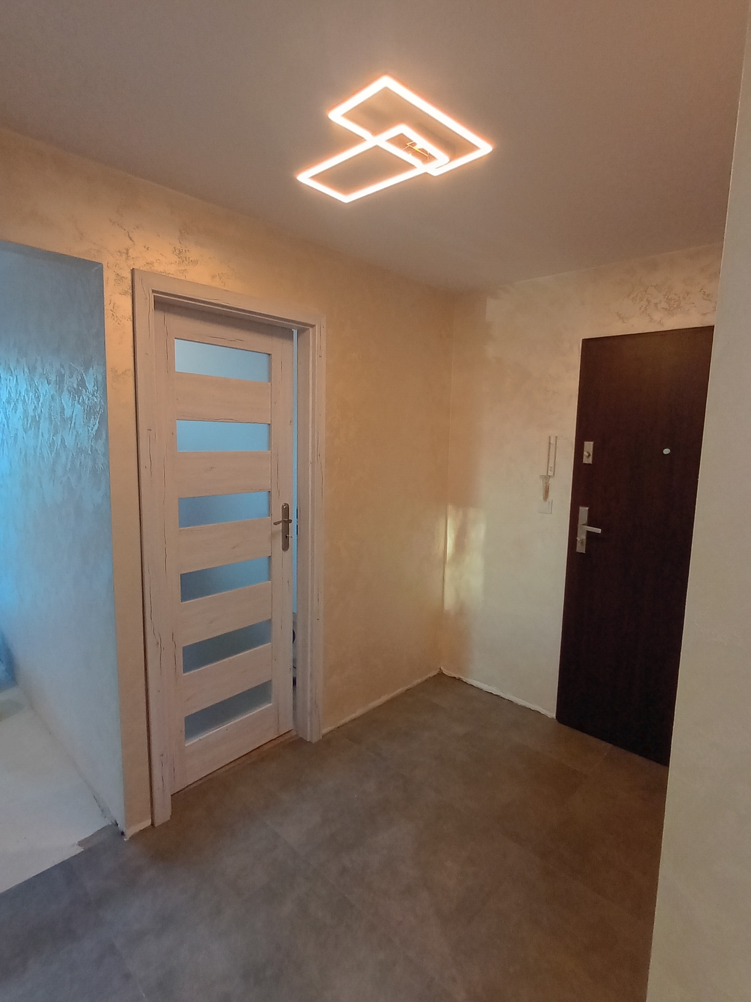 Wykończony korytarz z szarą podłogą, kremowymi ścianami z efektem dekoracyjnym i nowoczesną lampą sufitową. Widoczne dwie pary drzwi.