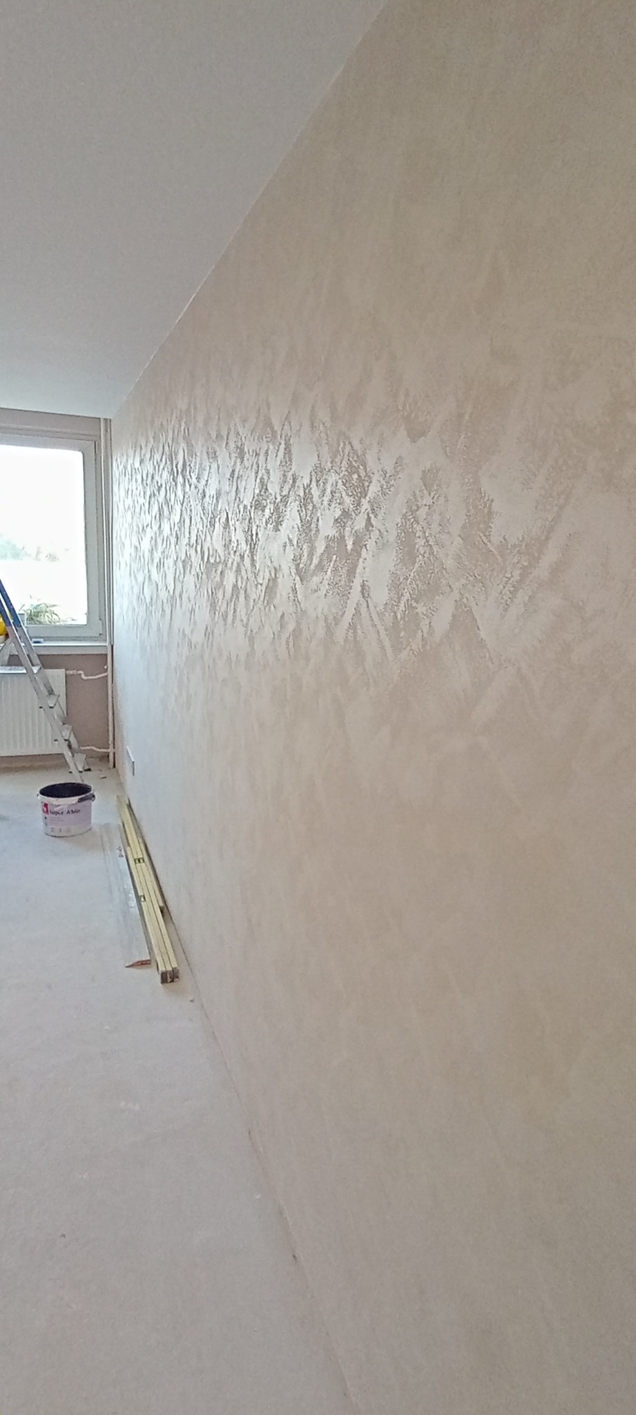 Ściana z dekoracyjną, beżową fakturą, widoczna z boku. Przy ścianie stoi drabina, puszka z farbą i listwy przypodłogowe. W tle okno.