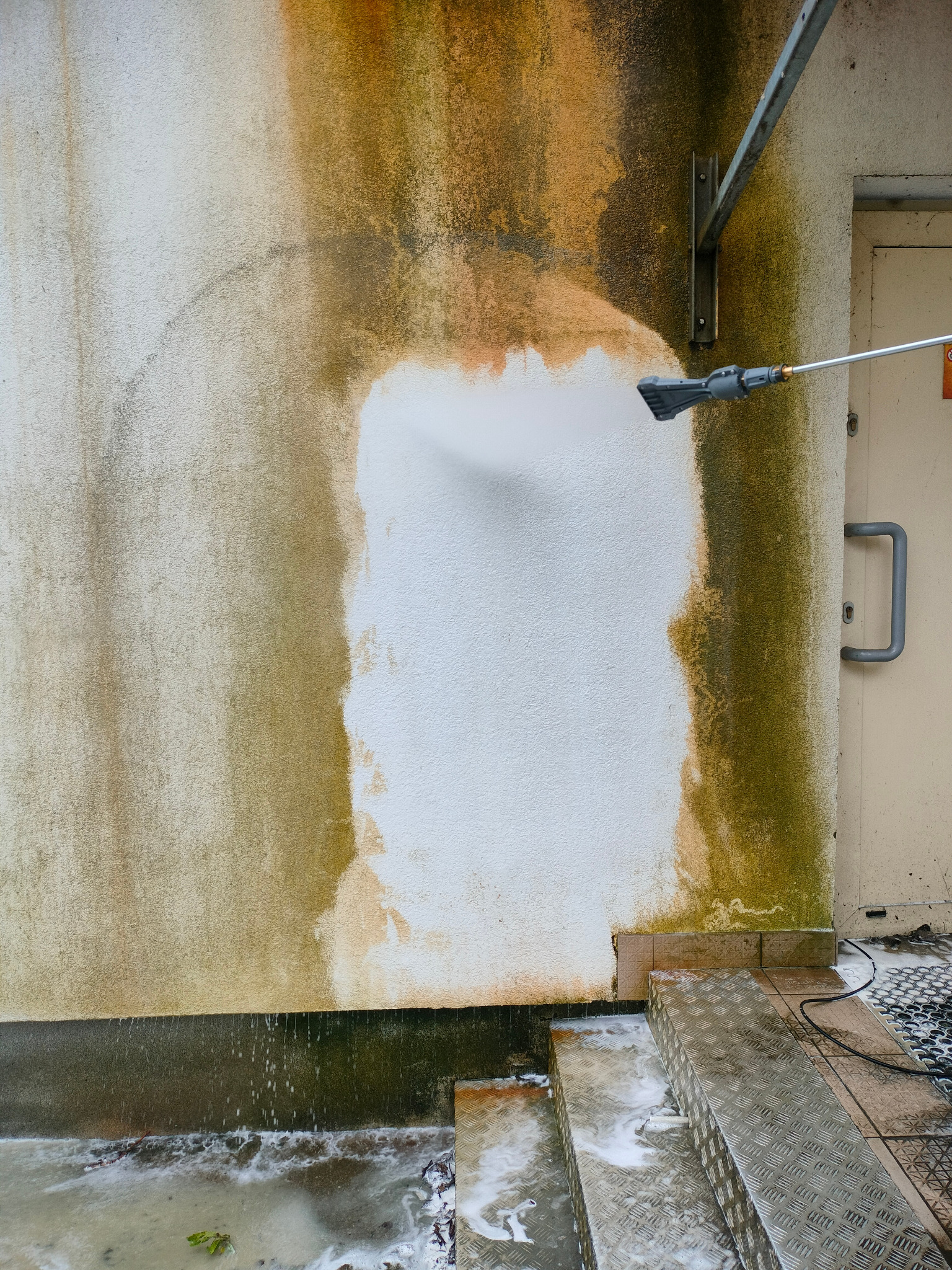 Mycie elewacji budynku z wyraźnymi zabrudzeniami, czyszczenie za pomocą myjki ciśnieniowej, widoczne efekty pracy, schody i fragment drzwi.