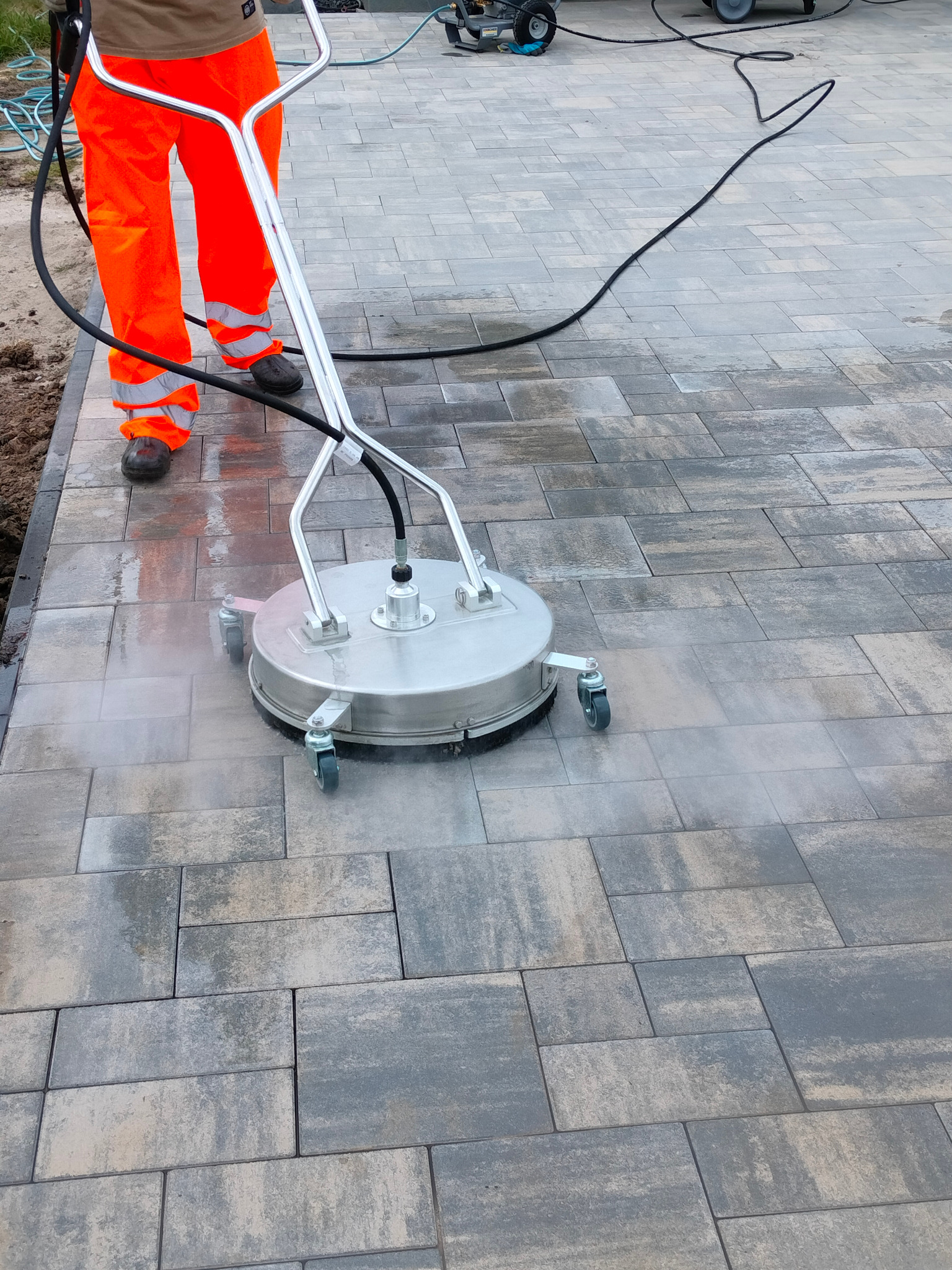 Czyszczenie kostki brukowej myjką ciśnieniową z obrotową głowicą. Operator w pomarańczowych spodniach roboczych. Widoczny efekt czyszczenia.