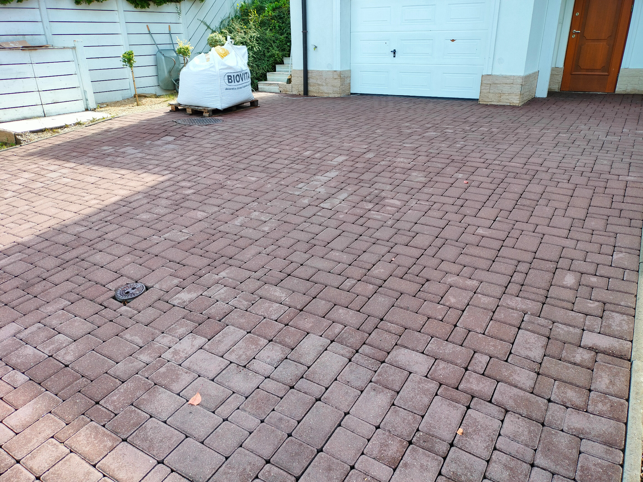 Ułożona kostka brukowa w odcieniach czerwieni na podjeździe. Widoczny właz kanalizacyjny i paleta z workiem. Brama garażowa i drzwi wejściowe w tle.