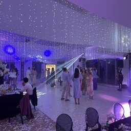 AGENCJA ARTYSTYCZNA MOSQUITO DARIUSZ ORŁOWSKI-TRYZNO - Uroczystość w sali z dekoracją świetlną: wiszące światła, reflektory, zespół muzyczny z saksofonem. Konfetti na podłodze, ustawione stoły z kwiatami.