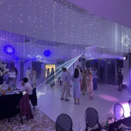 Uroczystość w sali z dekoracją świetlną: wiszące światła, reflektory, zespół muzyczny z saksofonem. Konfetti na podłodze, ustawione stoły z kwiatami.