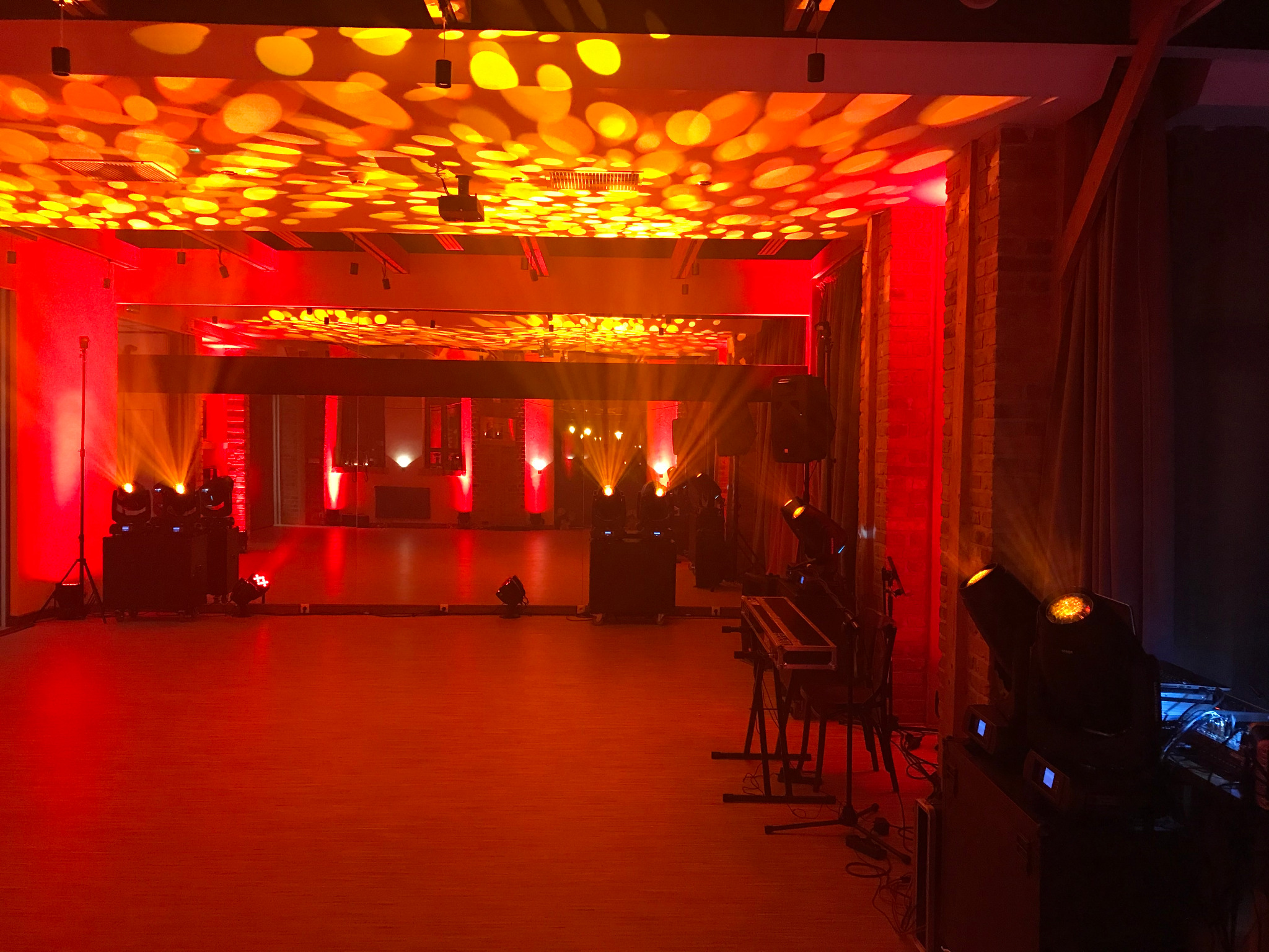 Sala z parkietem oświetlona czerwonym światłem i reflektorami. Na suficie widoczne wzory świetlne, tworzące klimat dyskoteki. Sprzęt nagłośnieniowy i klawisze w tle.