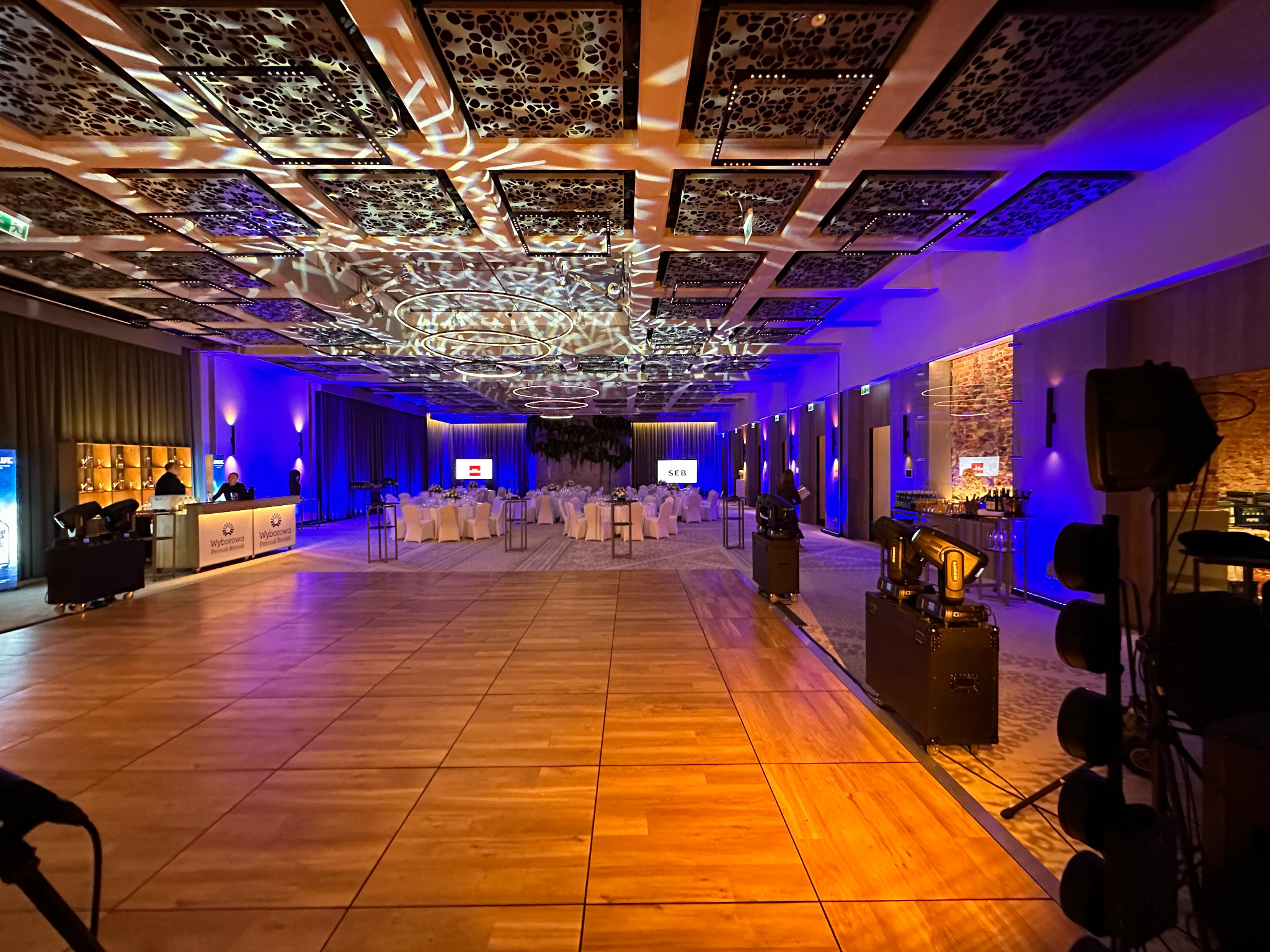 Sala bankietowa z parkietem, oświetleniem punktowym i dekoracyjnym na suficie. Widoczny barek, stoły z krzesłami przygotowane na uroczystość. Ustawione nagłośnienie.