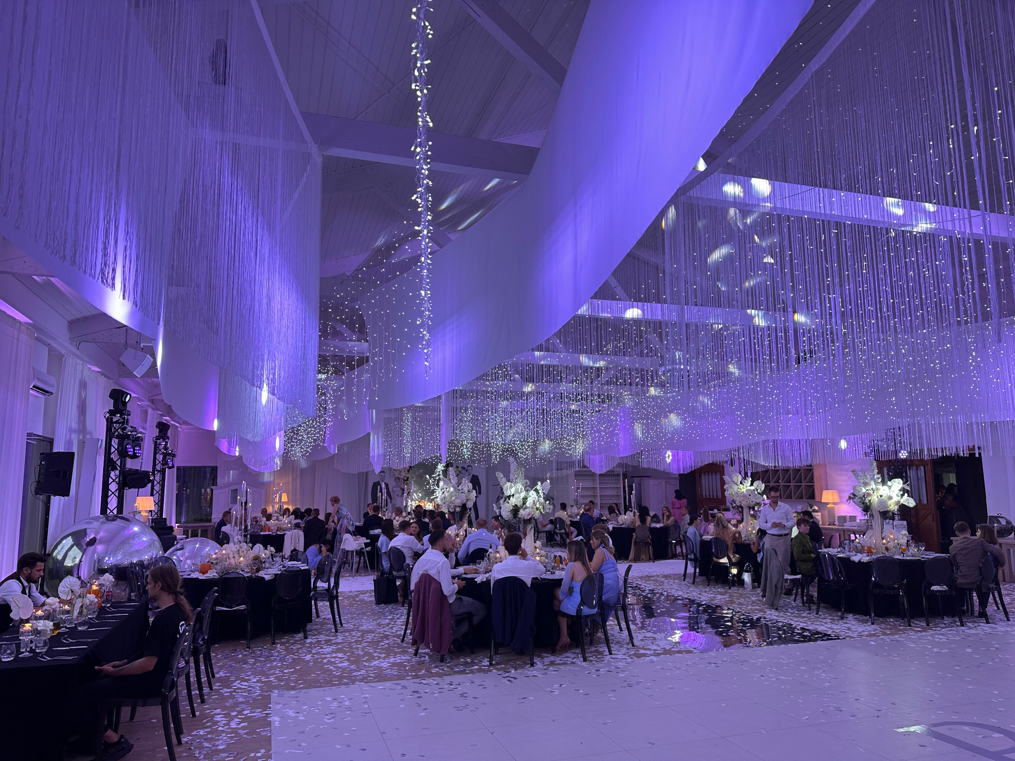 Udekorowana sala balowa z oświetleniem w odcieniach fioletu, wiszącymi dekoracjami i stolikami dla gości podczas przyjęcia.