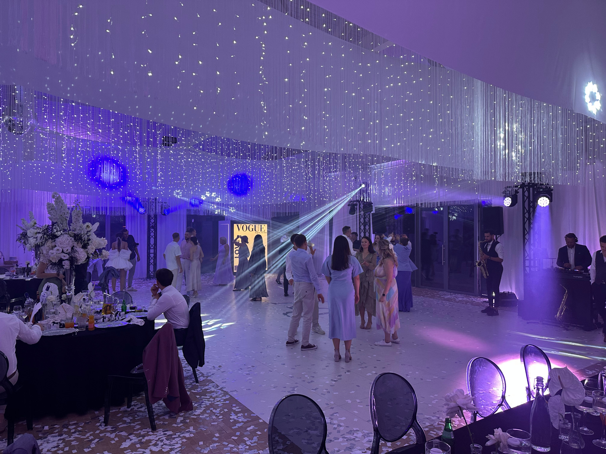 Uroczystość w sali z dekoracją świetlną: wiszące światła, reflektory, zespół muzyczny z saksofonem. Konfetti na podłodze, ustawione stoły z kwiatami.