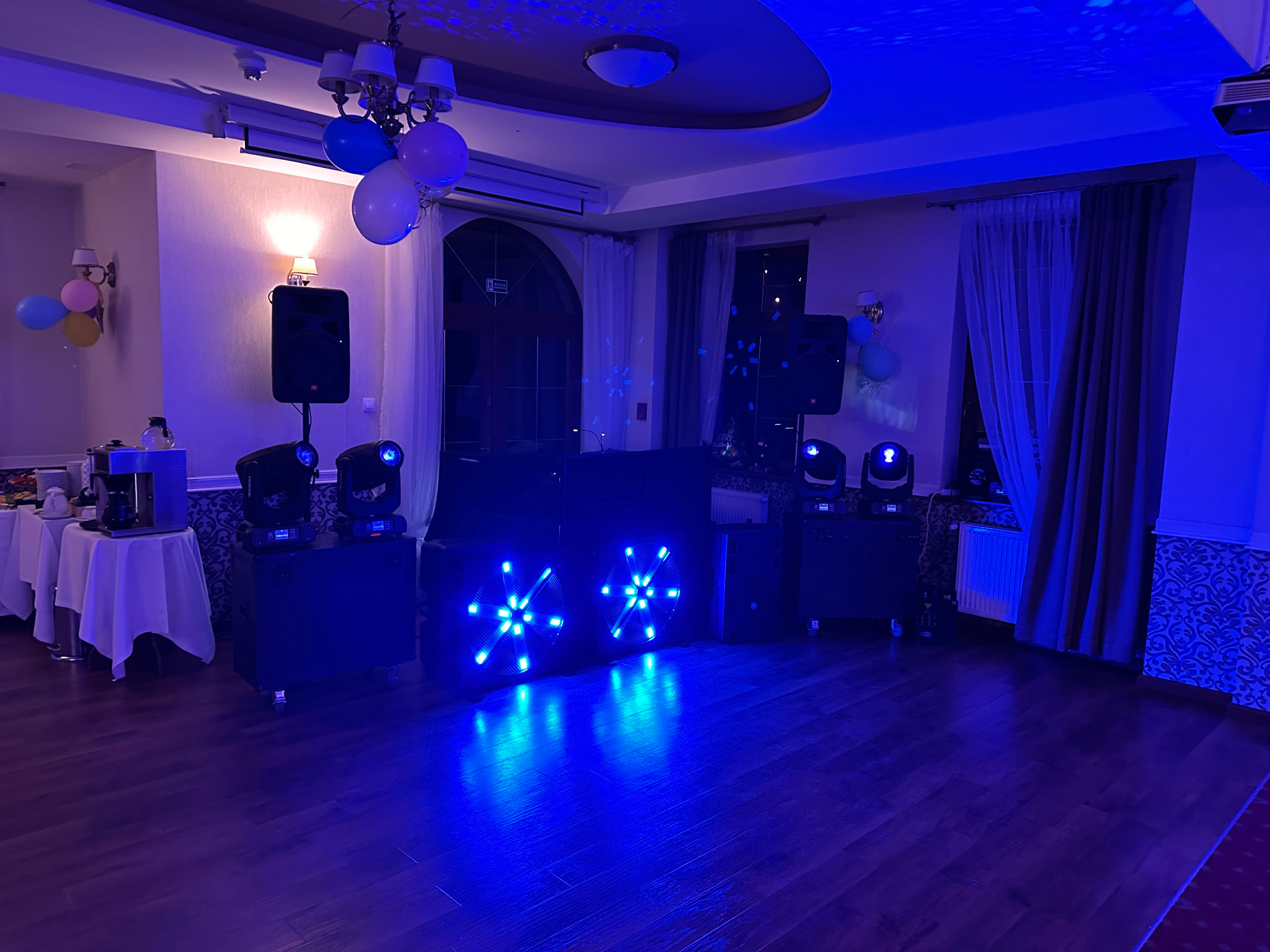 Sala bankietowa z nagłośnieniem i oświetleniem w niebieskiej tonacji. Widoczne głośniki, reflektory, konsoleta DJ-ska, dekoracje balonowe.