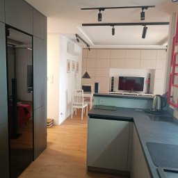 Aden - Nowoczesne wnętrze: jasna podłoga, szara zabudowa kuchenna z ciemnym blatem, oświetlenie punktowe i widok na salon z elementami dekoracyjnymi. Minimalistyczny design.