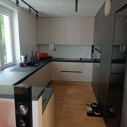 Aden - Nowoczesna kuchnia w jasnych barwach z drewnianą podłogą. Minimalistyczne szafki, czarny blat i oświetlenie szynowe. Miski dla zwierząt przy ścianie dodają domowego charakteru.