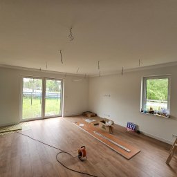 Aden - Jasne wnętrze w trakcie układania paneli podłogowych. Widoczne narzędzia, listwy przypodłogowe i drabina. Przez okno widać zieleń.