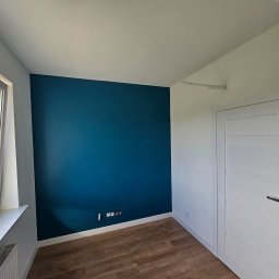 Aden - Pomalowane na turkusowo wnętrze pokoju z drewnianą podłogą, białymi ścianami i drzwiami. Widoczne gniazdka elektryczne i okno z widokiem na zieleń.